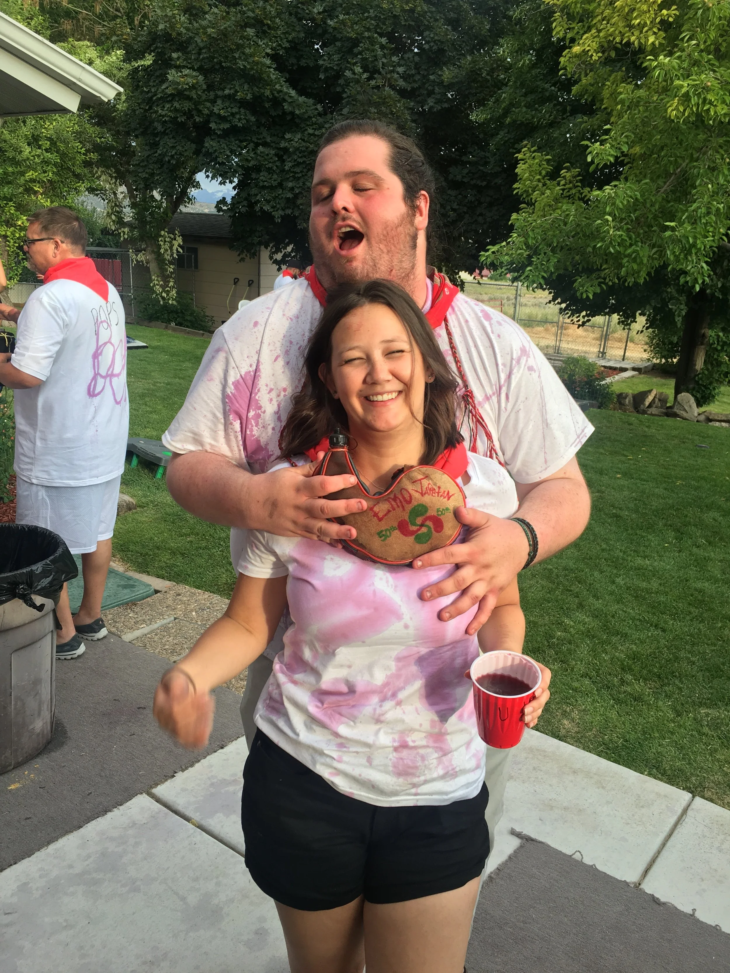A young woman smiling, holding a red cup, standing in front of a man, at an outdoor gathering during daytime. The man has a beard and long hair, and is wearing a white shirt with pink stains and a red bandana. The woman has long dark hair, and both are dressed casually in summer clothes. In the background, there are other people, trees, a house, and a yard.