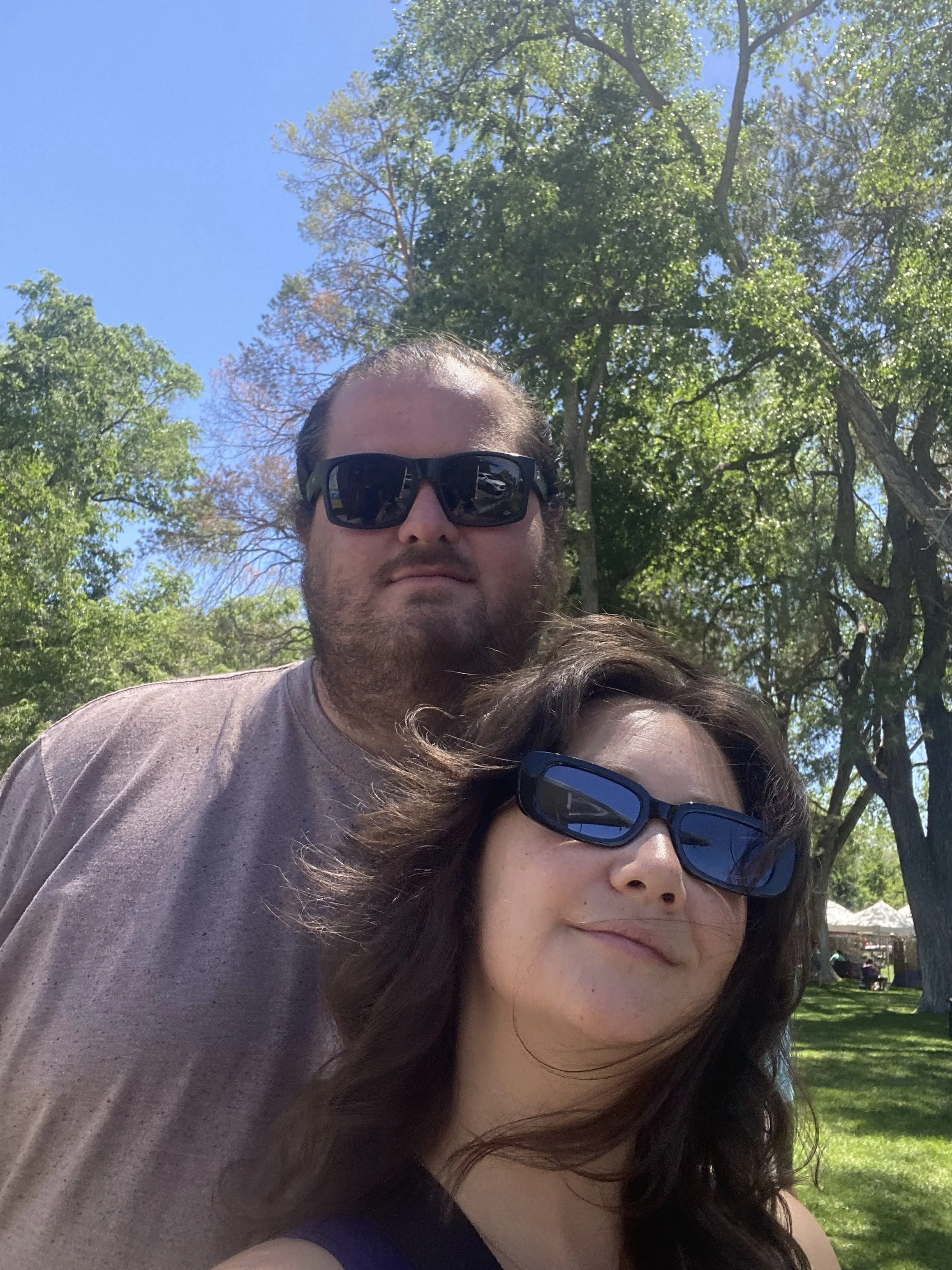 A man and a woman wearing sunglasses in a park with trees and blue sky in the background.