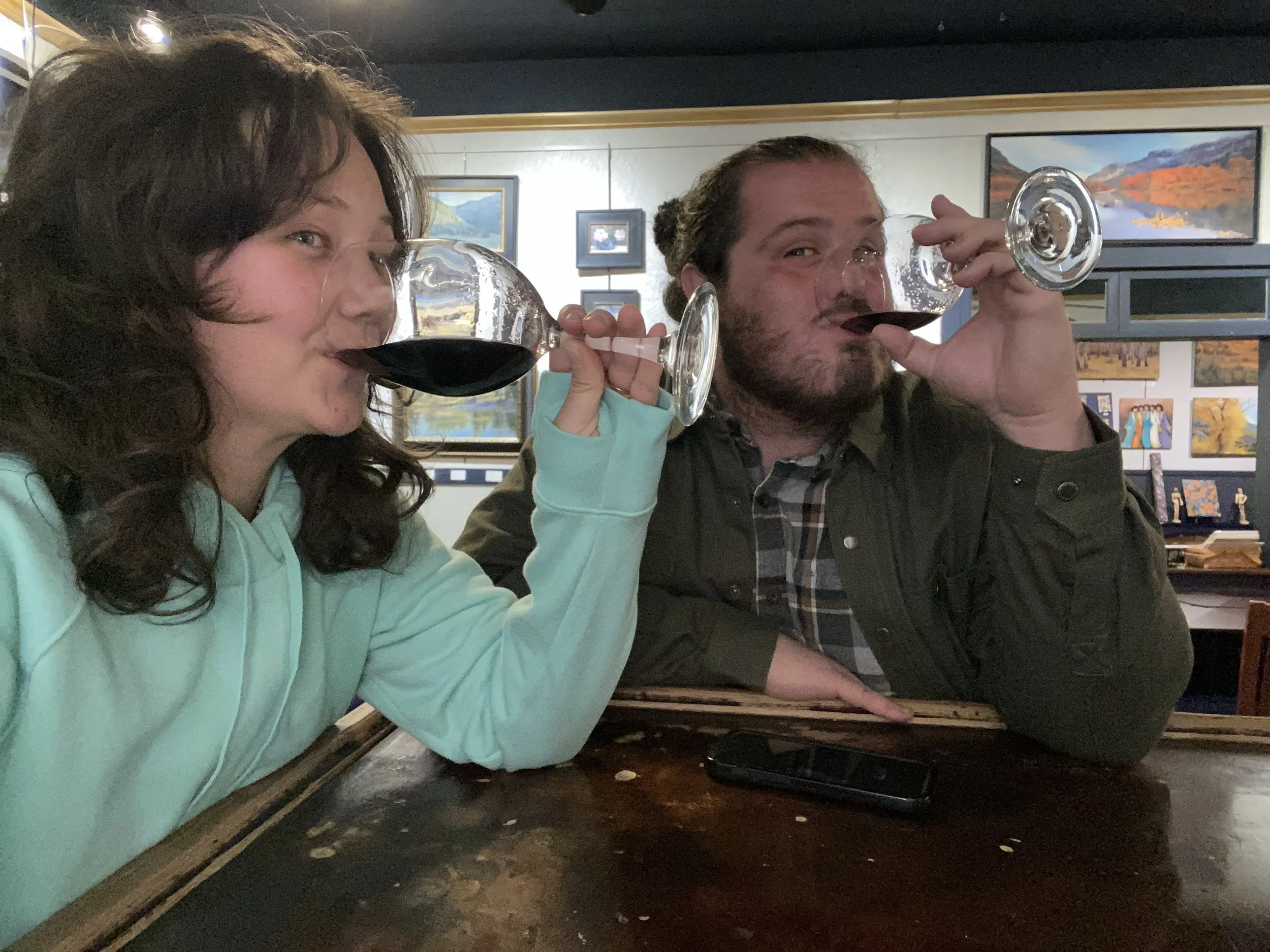 Two women are sitting at a table in an art gallery, drinking red wine from large wine glasses. They are smiling and enjoying their time together. The background shows framed landscape paintings on the wall.