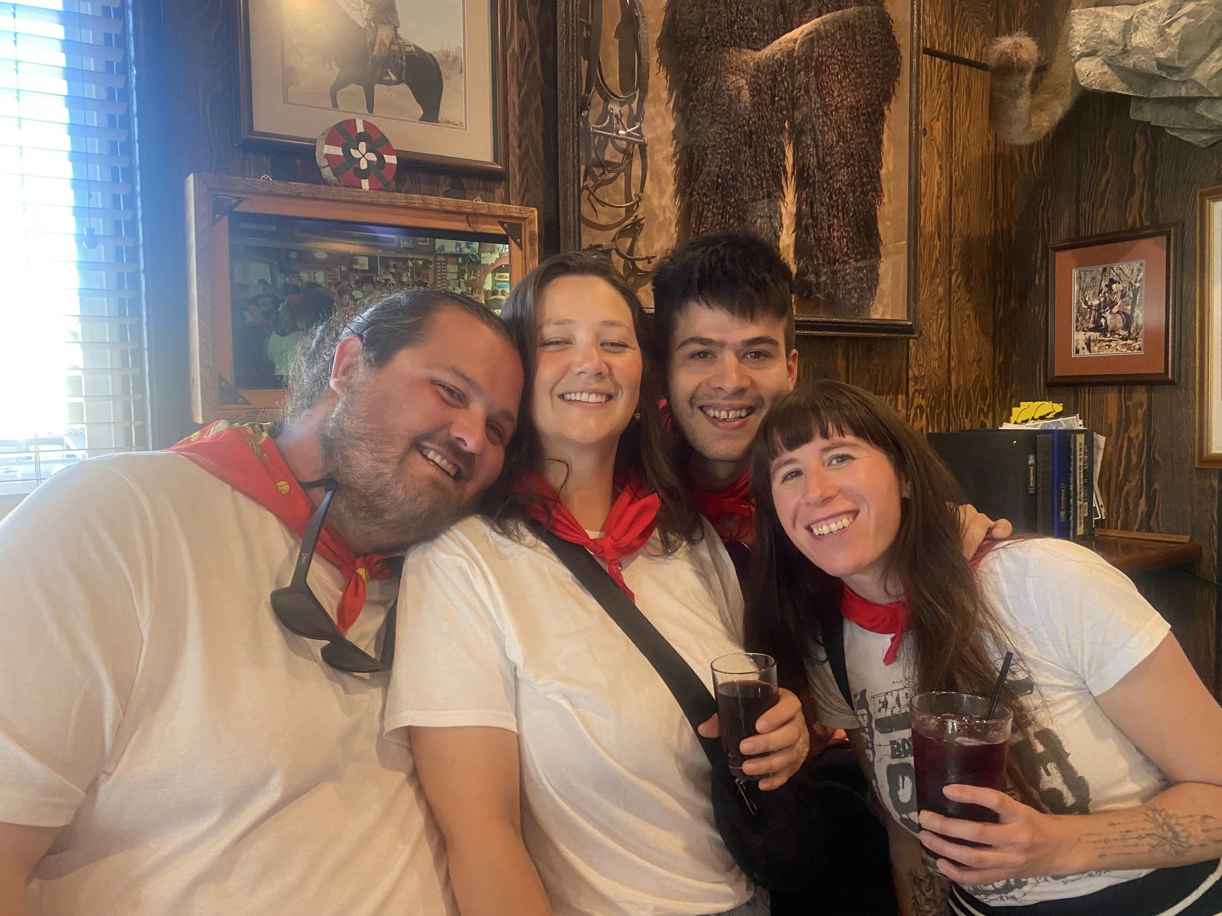A group of four people smiling and holding drinks at a restaurant or bar with a wooden wall and animal-themed decorations in the background.