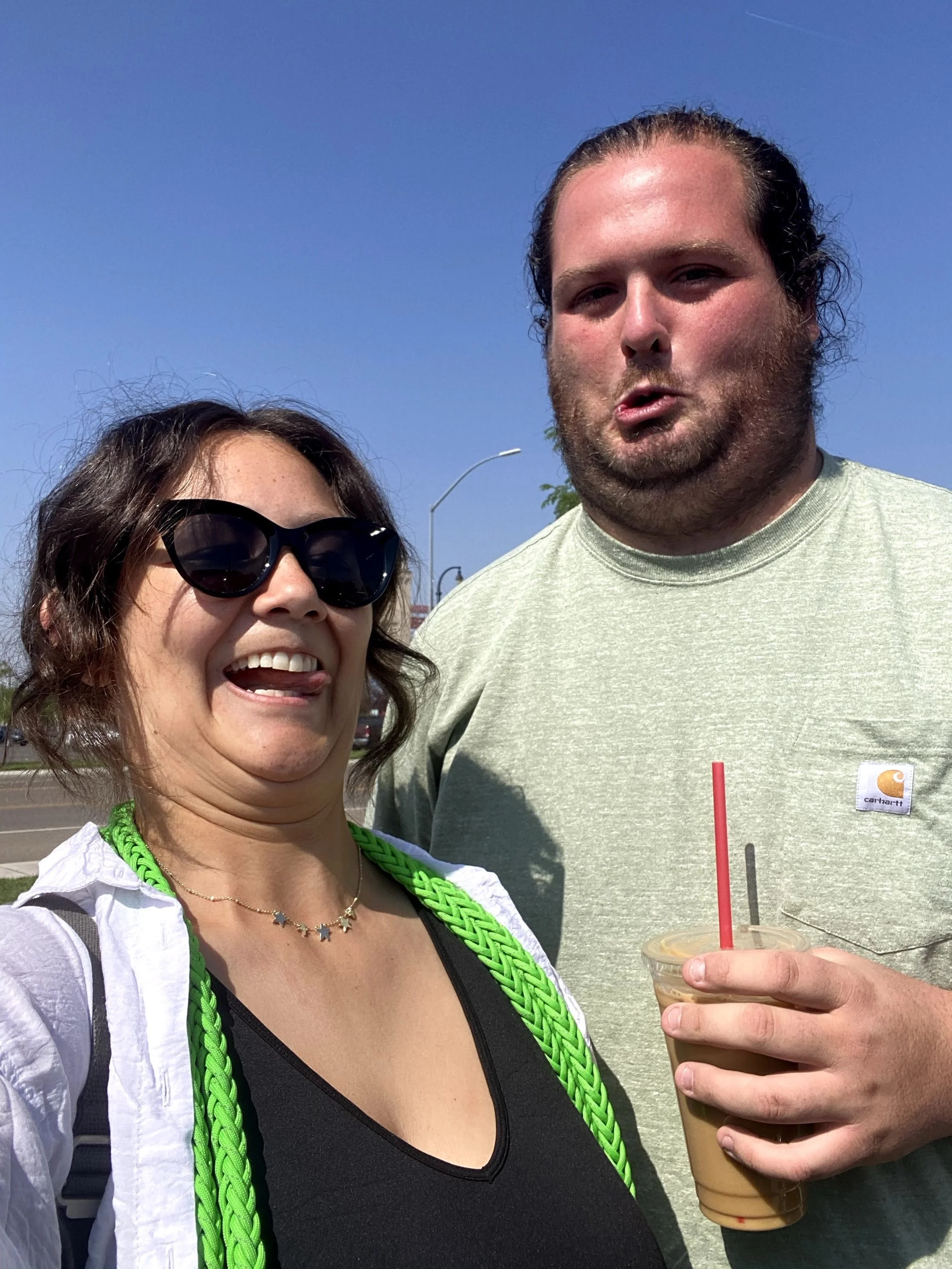 A woman wearing sunglasses, a necklace, and a green sunglasses strap is smiling and sticking out her tongue. A man next to her is holding a coffee drink with a pink straw and making a puckered face. They are outdoors on a sunny day.