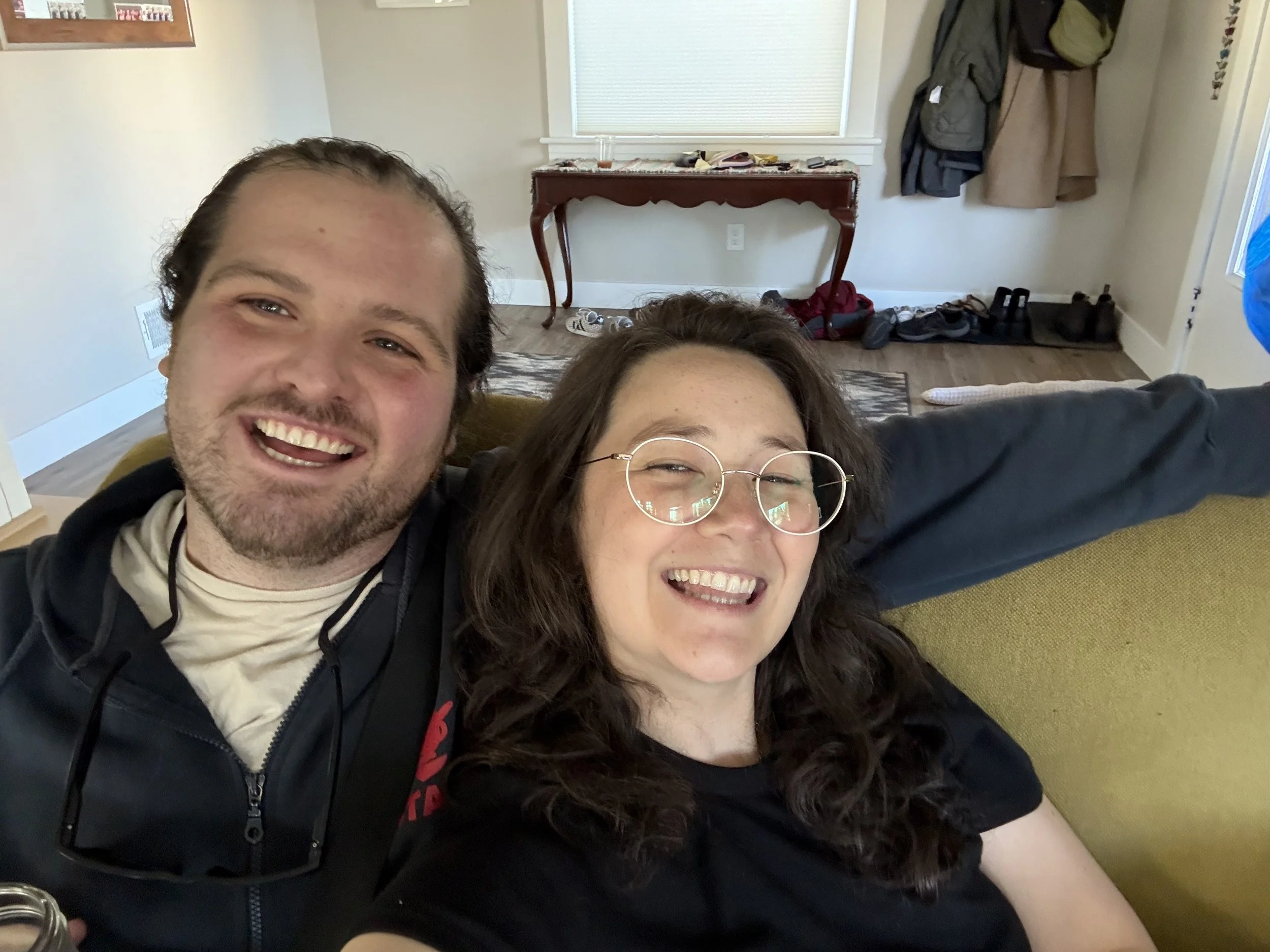 Close-up of a smiling man and woman sitting on a couch in a living room, surrounded by casual clothing and the interior of the room.