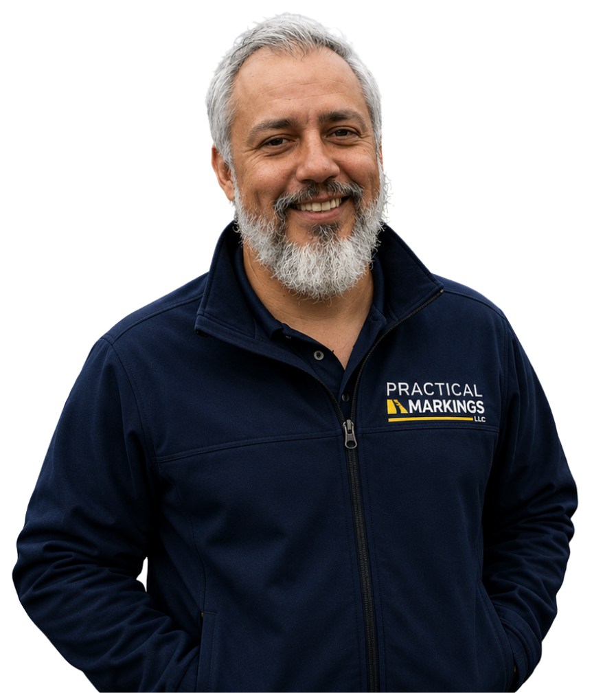A smiling man with gray hair and a beard wearing a navy blue zip-up jacket with the logo 'Practical Markings LLC' on it.