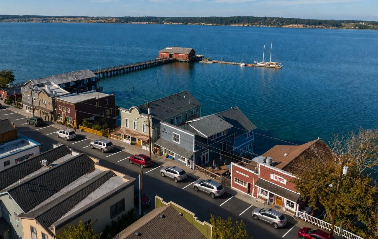 Aerial view of a seaside town featuring colorful buildings along a street, a waterway with a long pier extending into the water, and a small boat docked at the pier, with a body of water in the background.