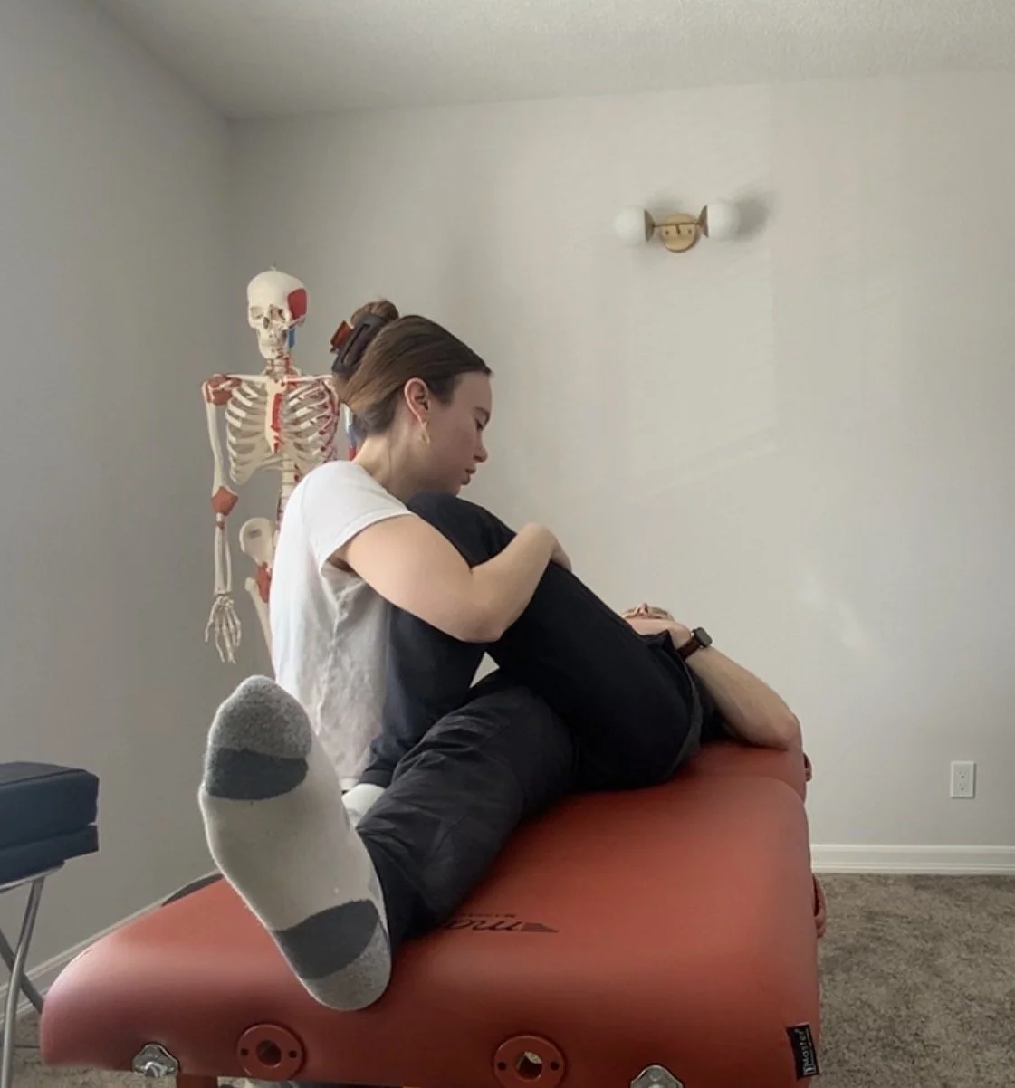 A woman providing osteopathic treatment to a man lying on an orange therapy table, with a skeleton model in the background and a wall light fixture on a plain white wall.