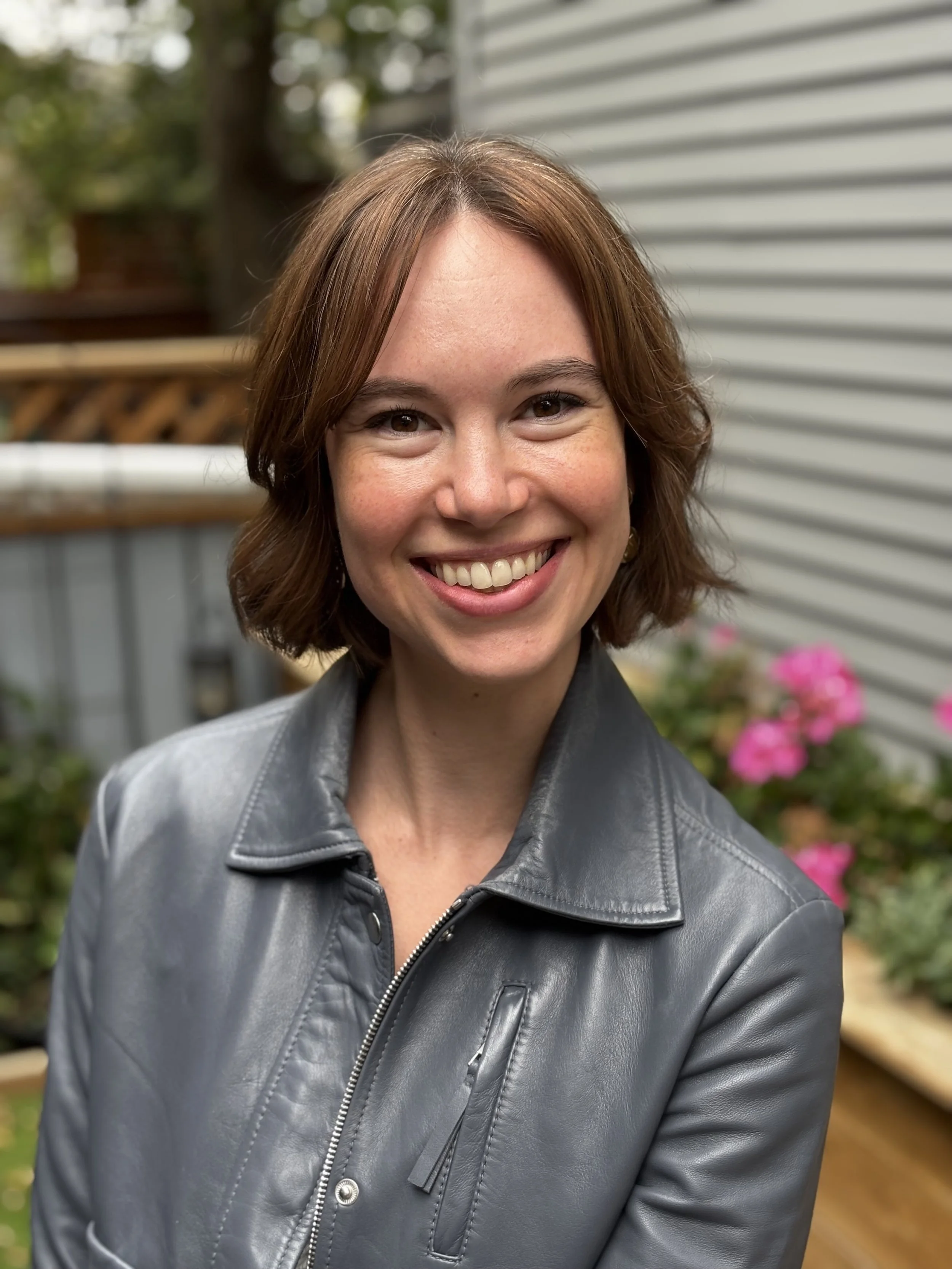 Close-up of a smiling woman with short brown hair wearing a gray leather jacket standing outdoors in front of a house with a gray siding wall and pink flowers.