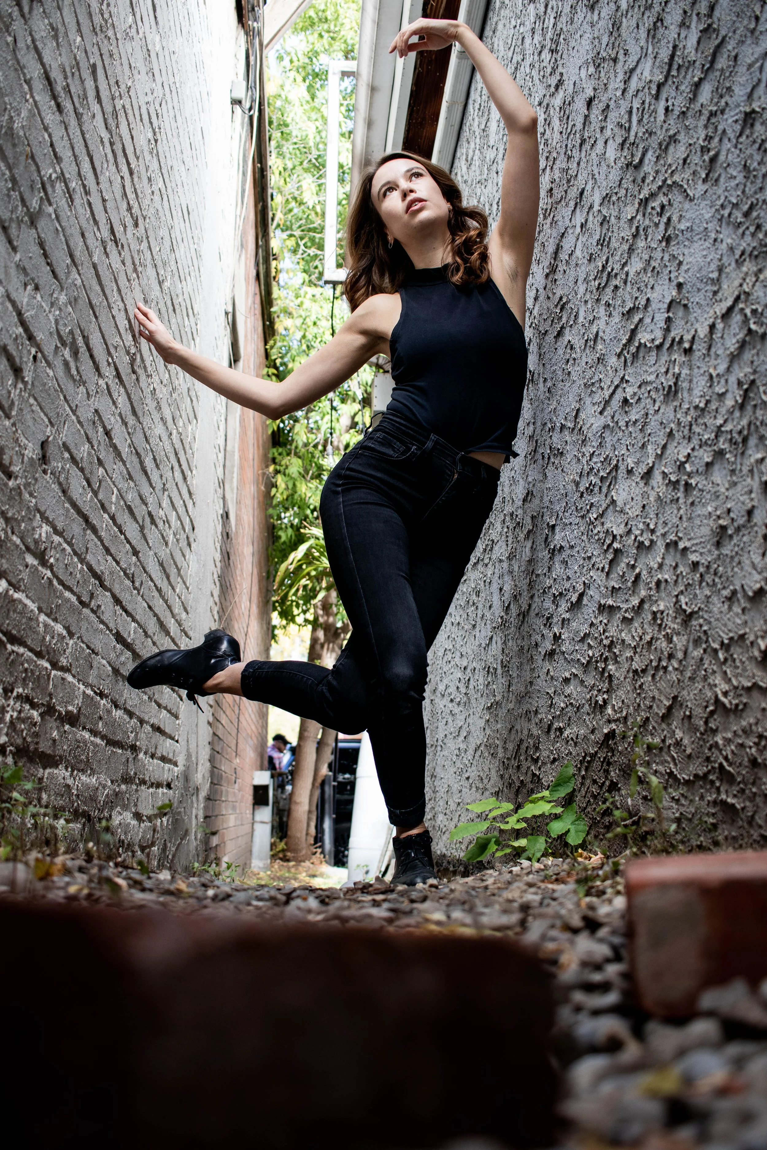 A woman with curly brown hair wearing a black sleeveless shirt and black pants is balancing on her right foot with her left leg raised, leaning against a textured brick wall in a narrow alleyway, with her right arm extended upward and her left hand holding the wall, near some plants and fallen leaves on the ground.