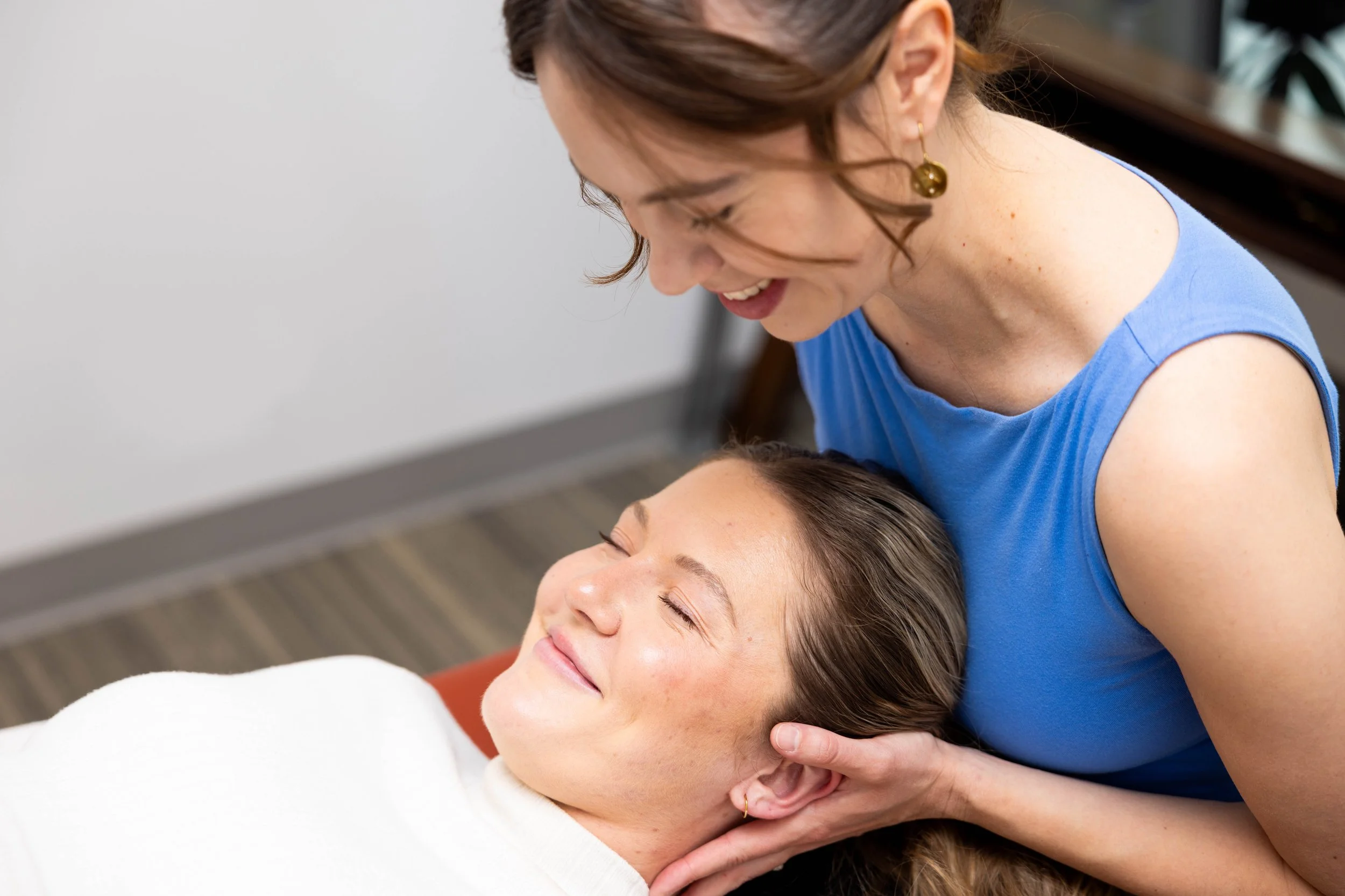 A woman applying osteopathic treatment to the patient’s neck. The patient is a women laying on her back on a treatment table. Both women are smiling, in a room with a wooden floor and shelves in the background.