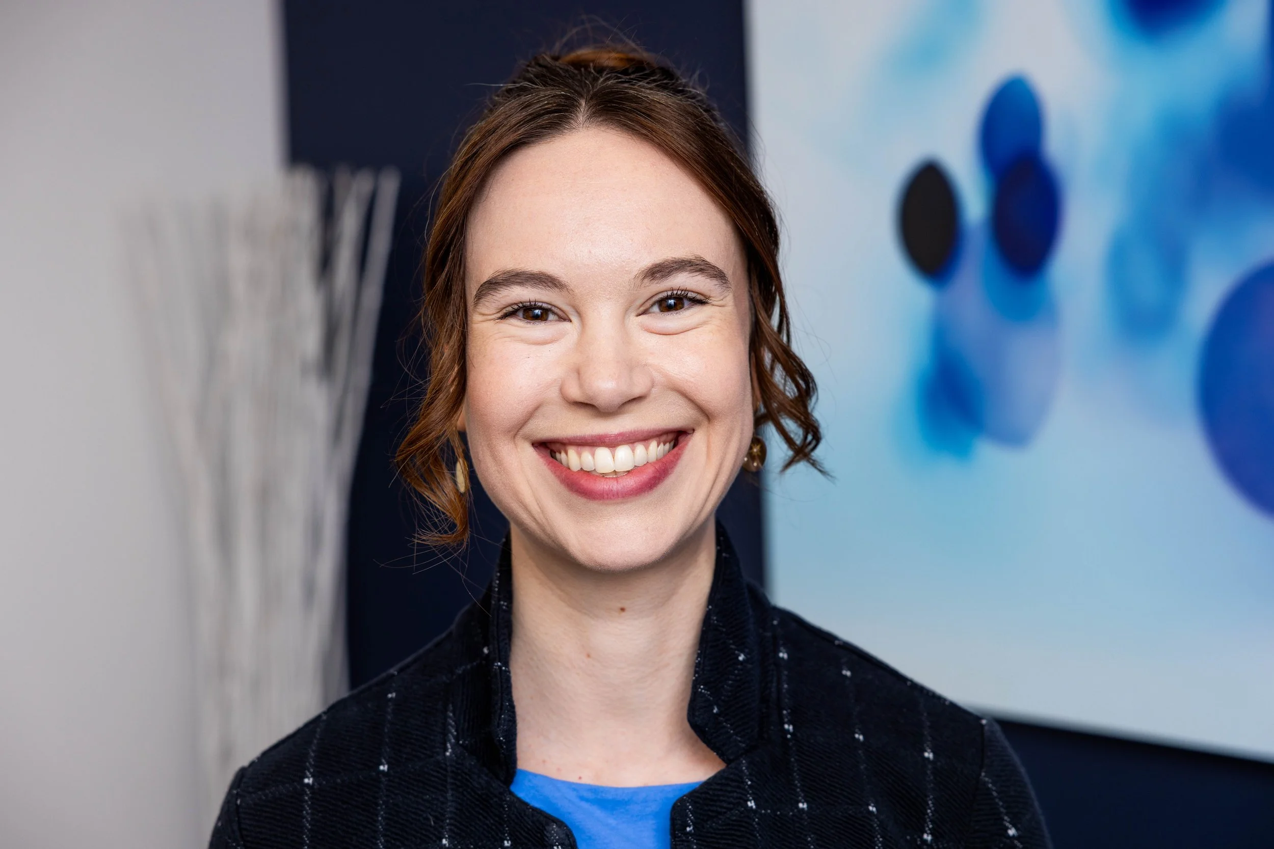 A smiling woman with short brown hair, wearing a black blazer with a subtle checked pattern and a blue top, standing in front of a blue abstract background with circular designs.