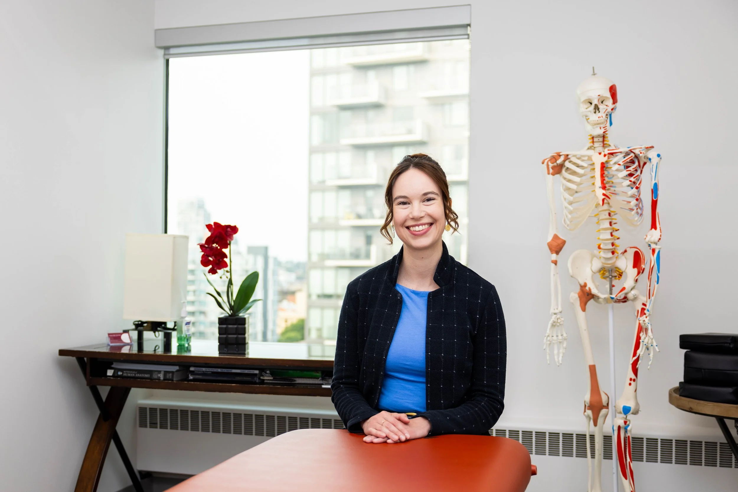 A young woman sitting at a table in an office or medical setting, smiling. There is a human skeleton model and a window with a city view in the background.
