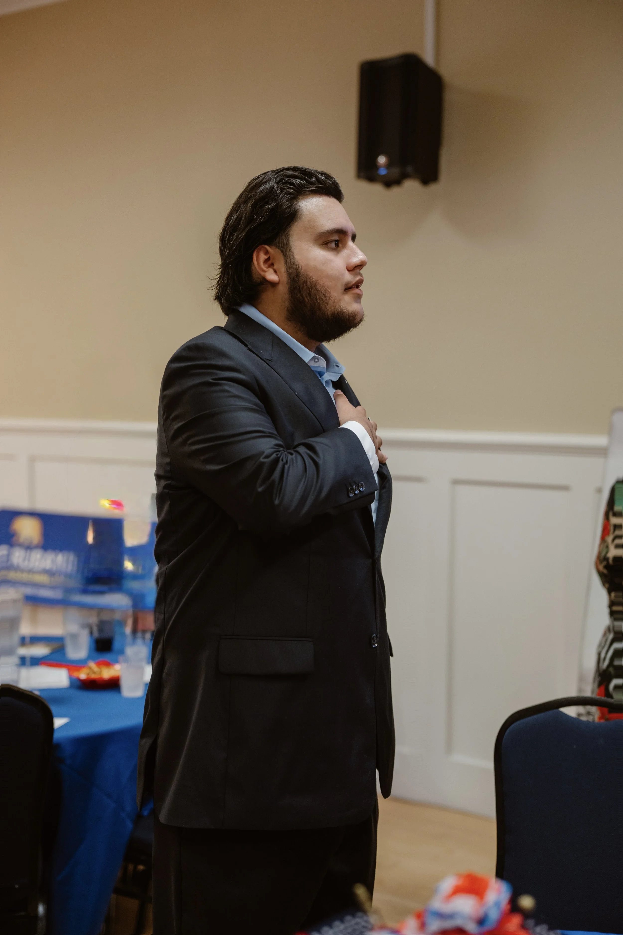 A man in a suit standing indoors with his hand over his heart, possibly during a patriotic moment or ceremony.
