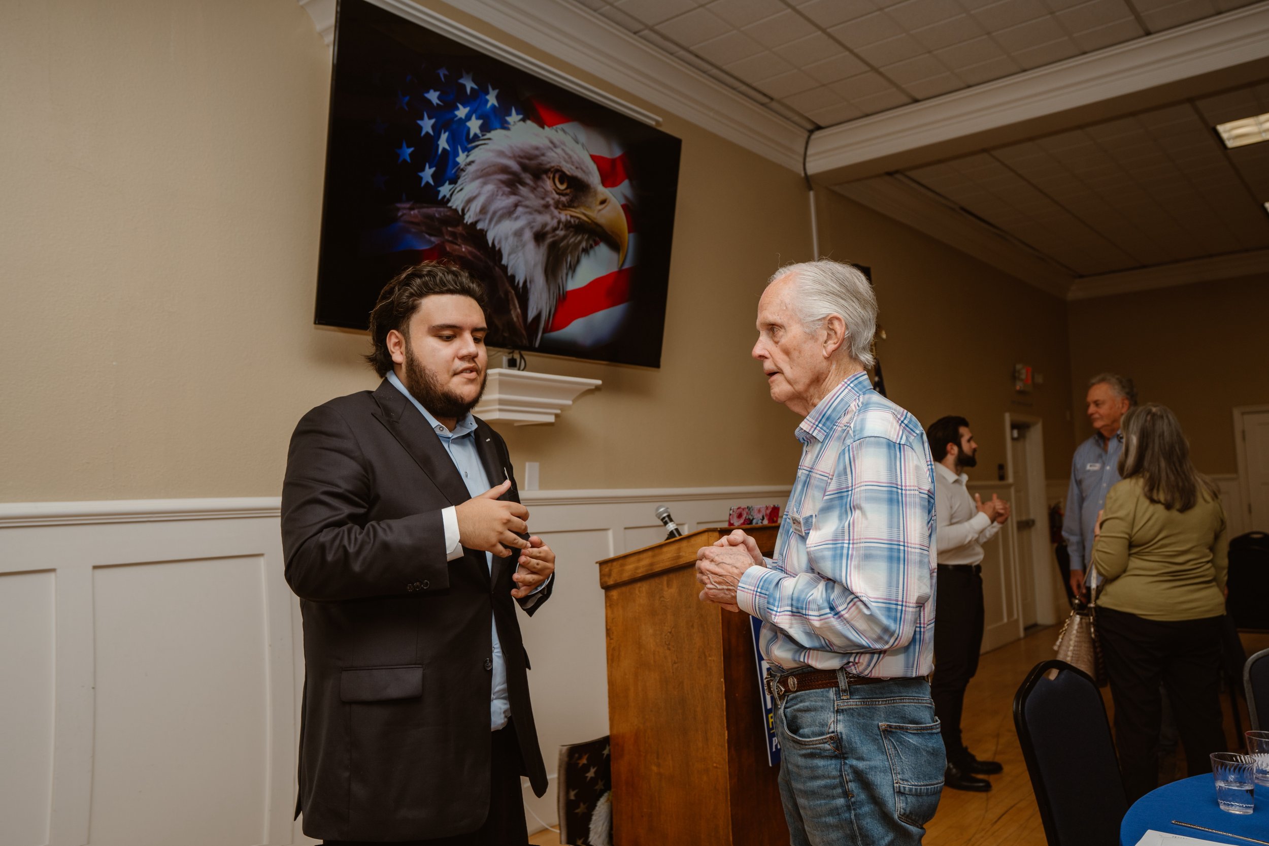An elderly man and a younger man in a suit engaging in conversation at a formal event in a room decorated with patriotic American symbols, including a painting of a bald eagle with the American flag in the background.