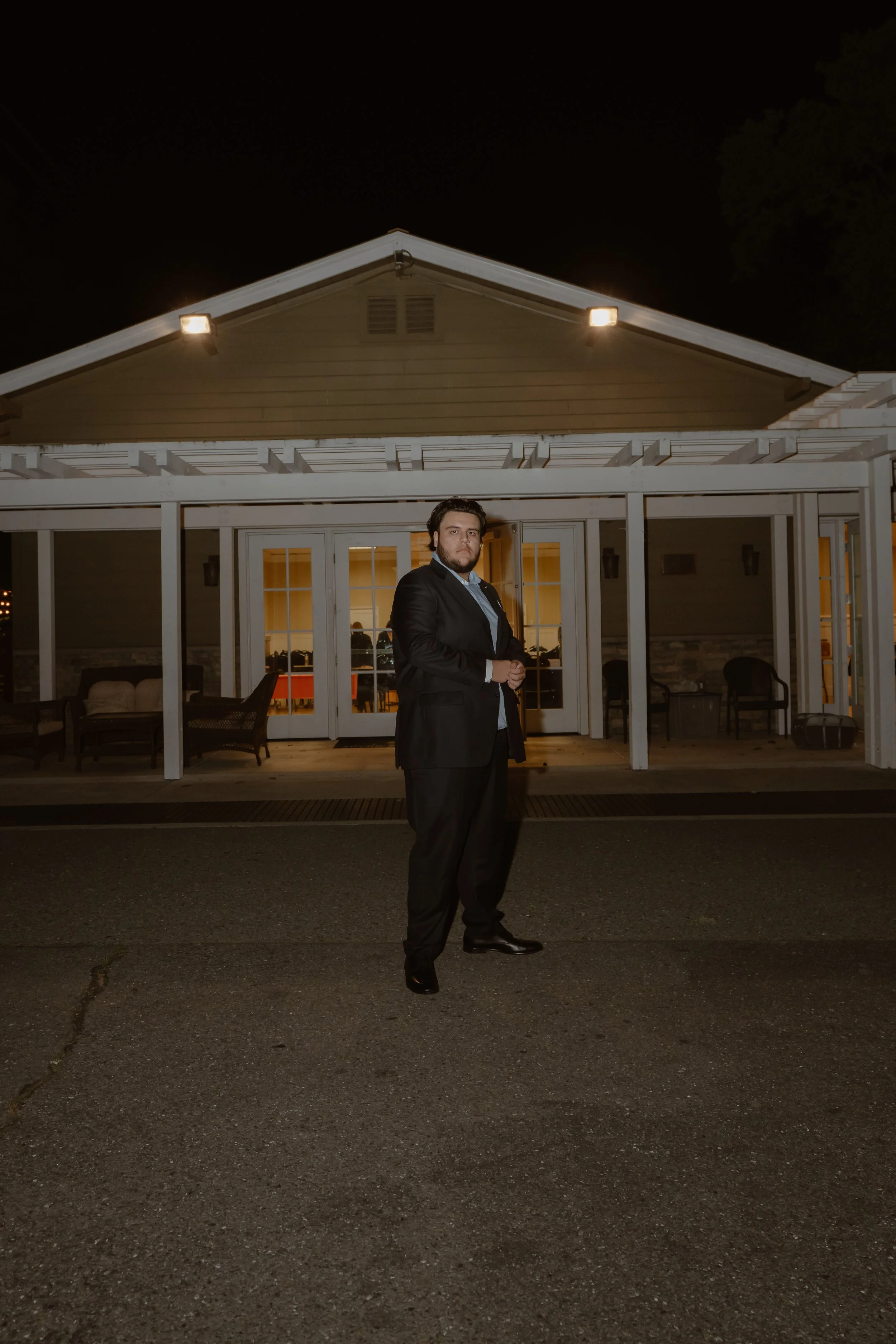 A man in a black suit standing outside at night in front of a house with lit windows and a porch. The house has a gabled roof and a glass door. The man has dark hair and a beard, and is standing with his hands clasped in front of him, looking into the camera.
