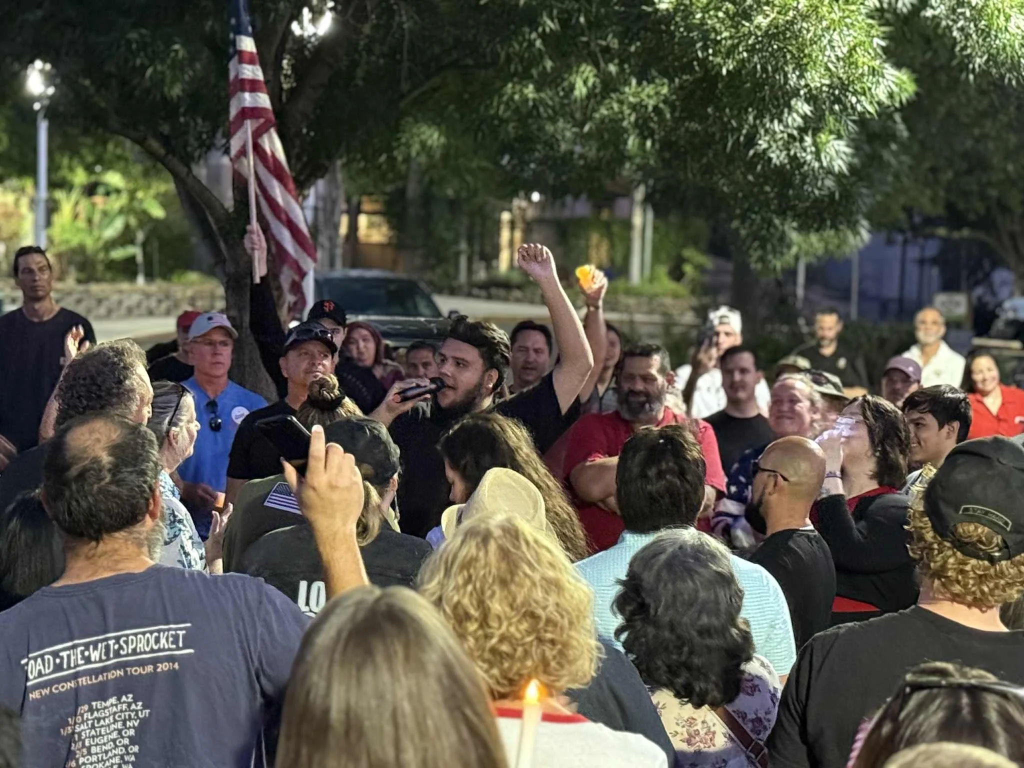 A diverse group of people gathered outdoors under trees, with someone at the center speaking into a microphone and raising a fist, while holding an American flag.