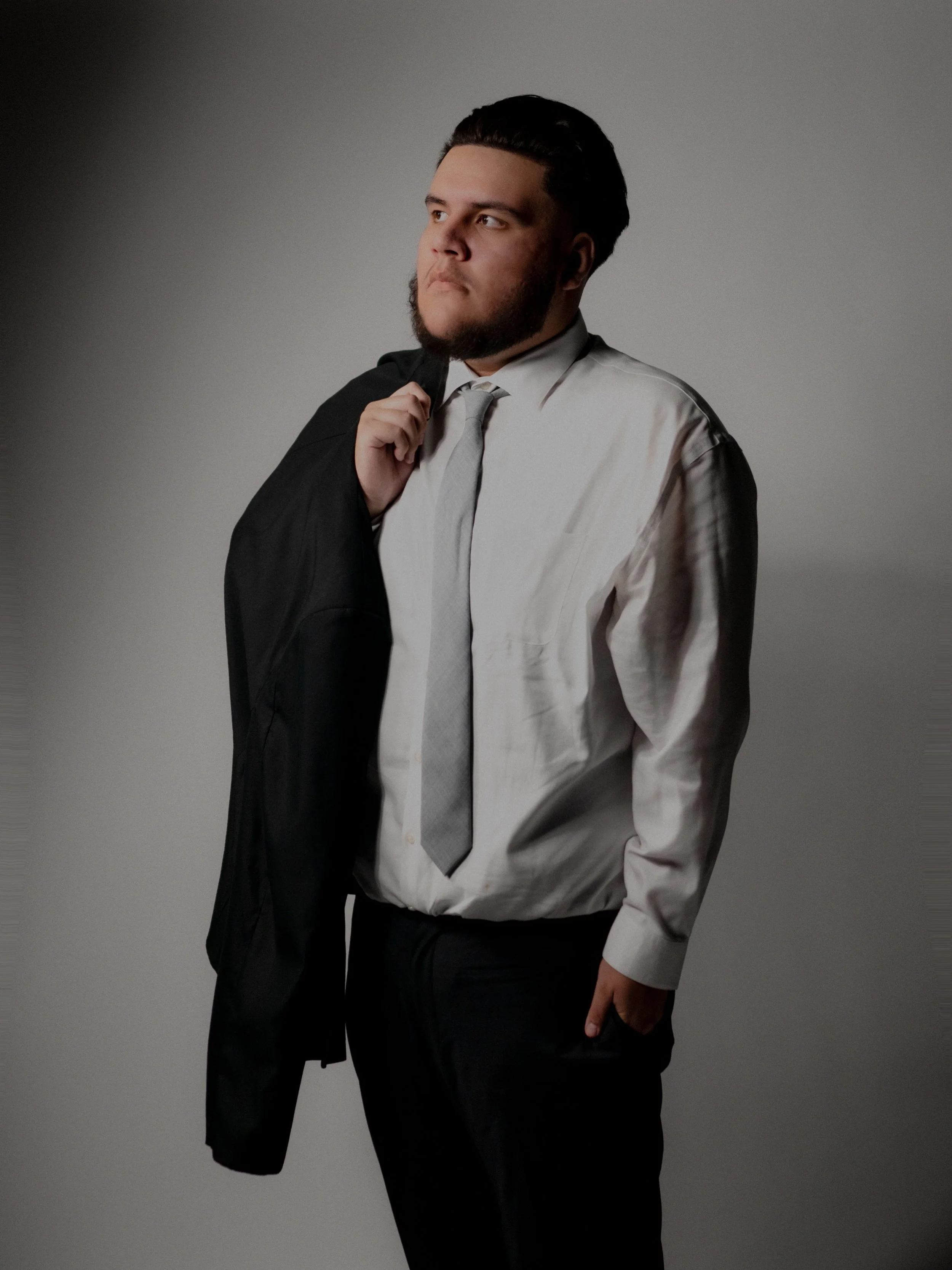 Man with a beard and slicked-back hair holding a suit jacket over his shoulder, wearing a white dress shirt and tie, posing against a plain gray background.