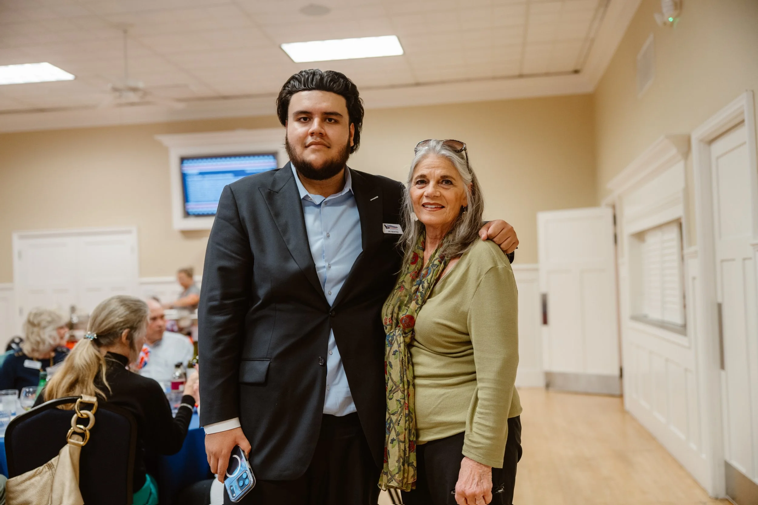 A young man in a suit and an older woman in a green top pose together at an indoor social gathering, smiling. The background shows people seated at tables and a wall-mounted monitor in a beige-colored room.