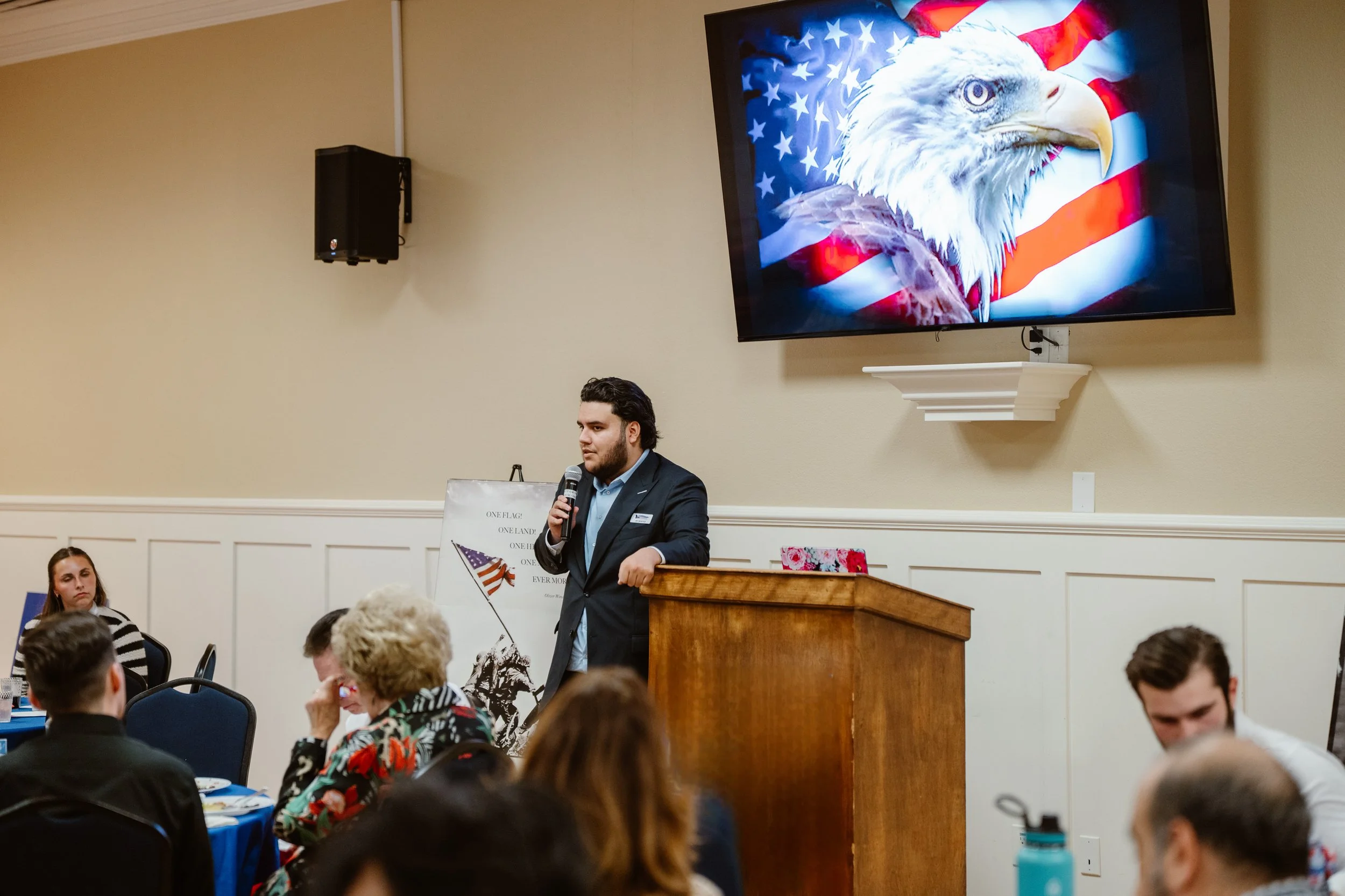 A man in a dark suit speaking at a podium in a room filled with people. Behind him is a large screen displaying an American flag and a bald eagle. Some attendees are sitting and listening, including a woman with a floral jacket.