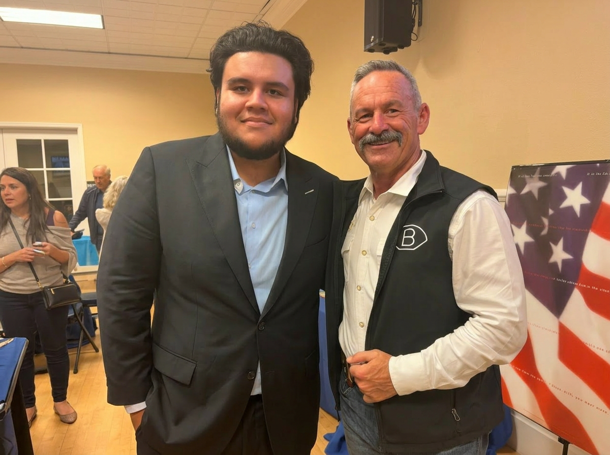 Two men standing together at an indoor event, smiling at the camera. The man on the left is younger, with dark hair and a beard, wearing a gray suit and light blue shirt. The man on the right is older, with gray hair and mustache, wearing a black vest over a white shirt. In the background, there are other people and a flag with stars and stripes partially visible.