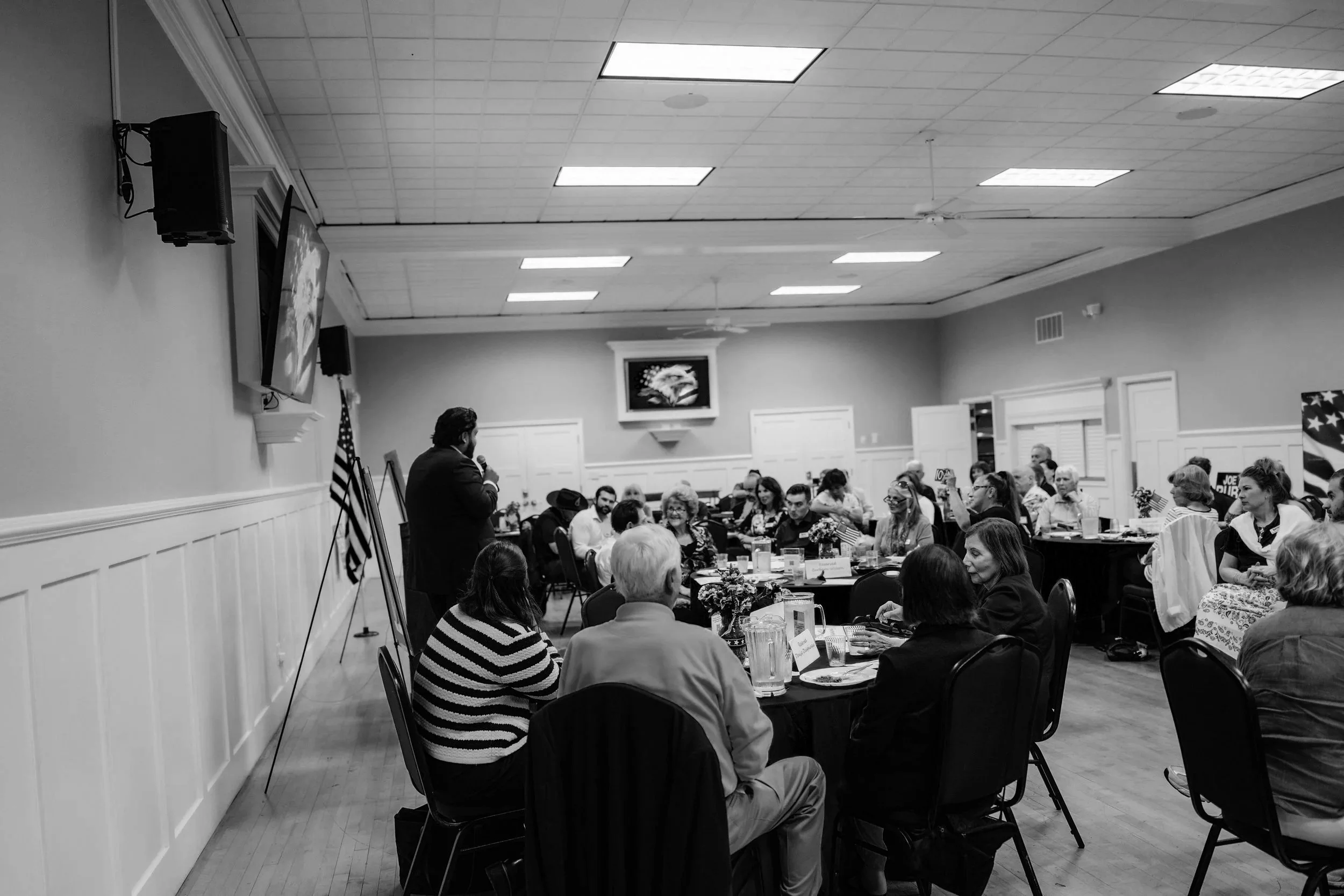 A man in a suit holding a microphone speaking to a seated audience in a banquet room. The audience is diverse, sitting at round tables with floral centerpieces, and appears to be engaged in the event. The room features a large flat-screen TV, American flags, and ceiling fans.
