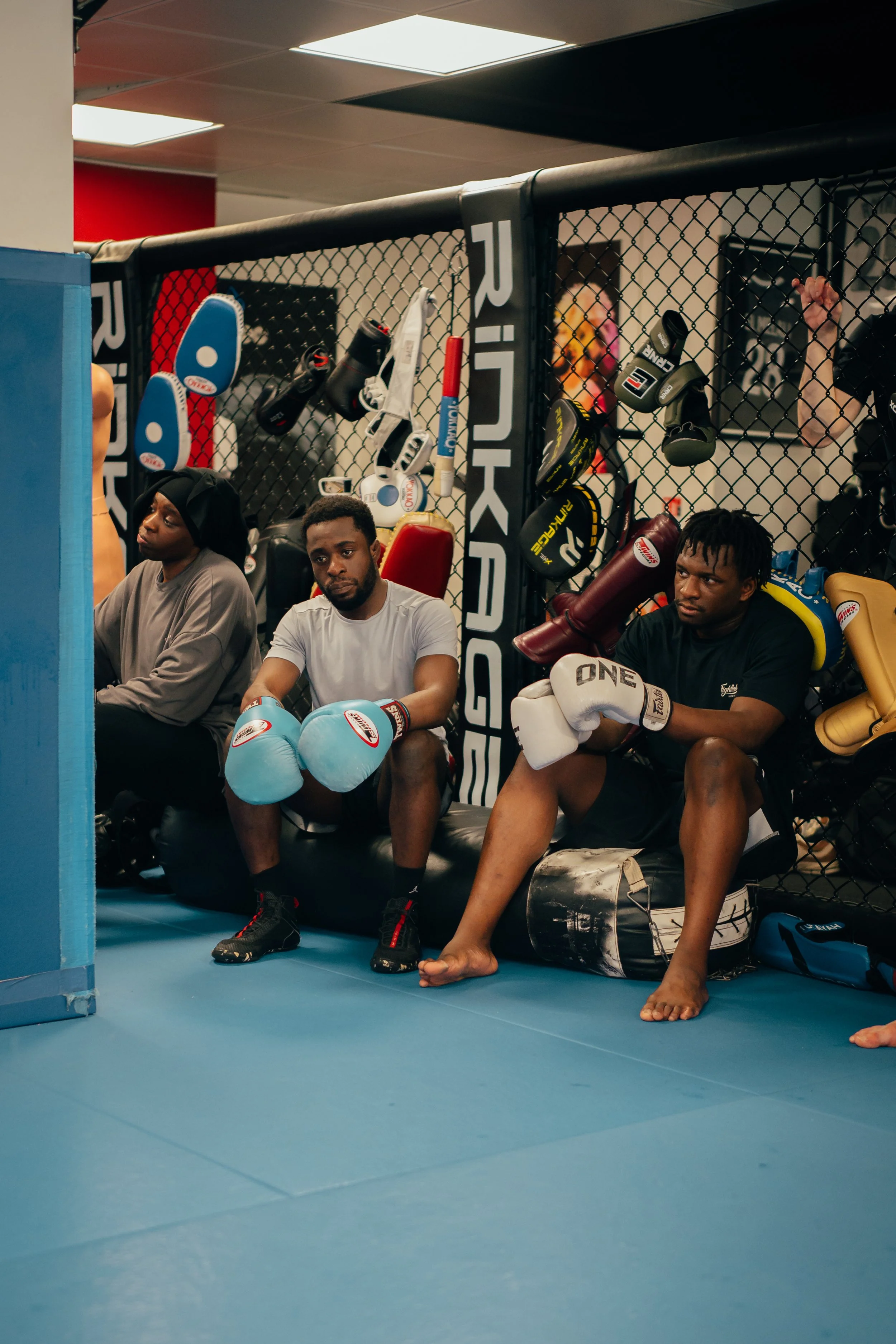 Trois hommes assis dans une cage de boxe, portant des gants de boxe, avec des sacs de sport suspendus derrière eux. Les hommes semblent fatigués ou concentrés, dans une ambiance de vestiaire ou de préparation à l'entraînement.