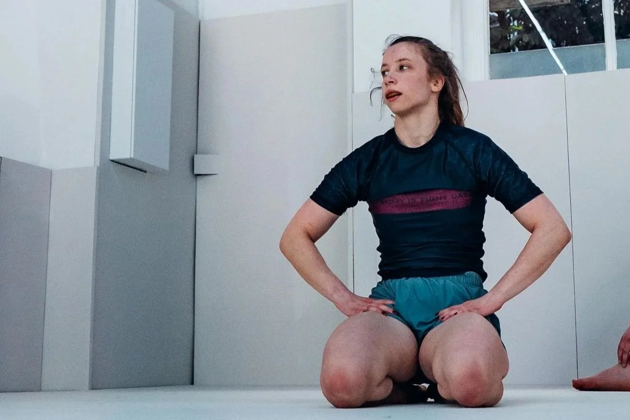 Jeune fille en tenue sportive, à genoux par terre, dans un dojo aux murs blancs.