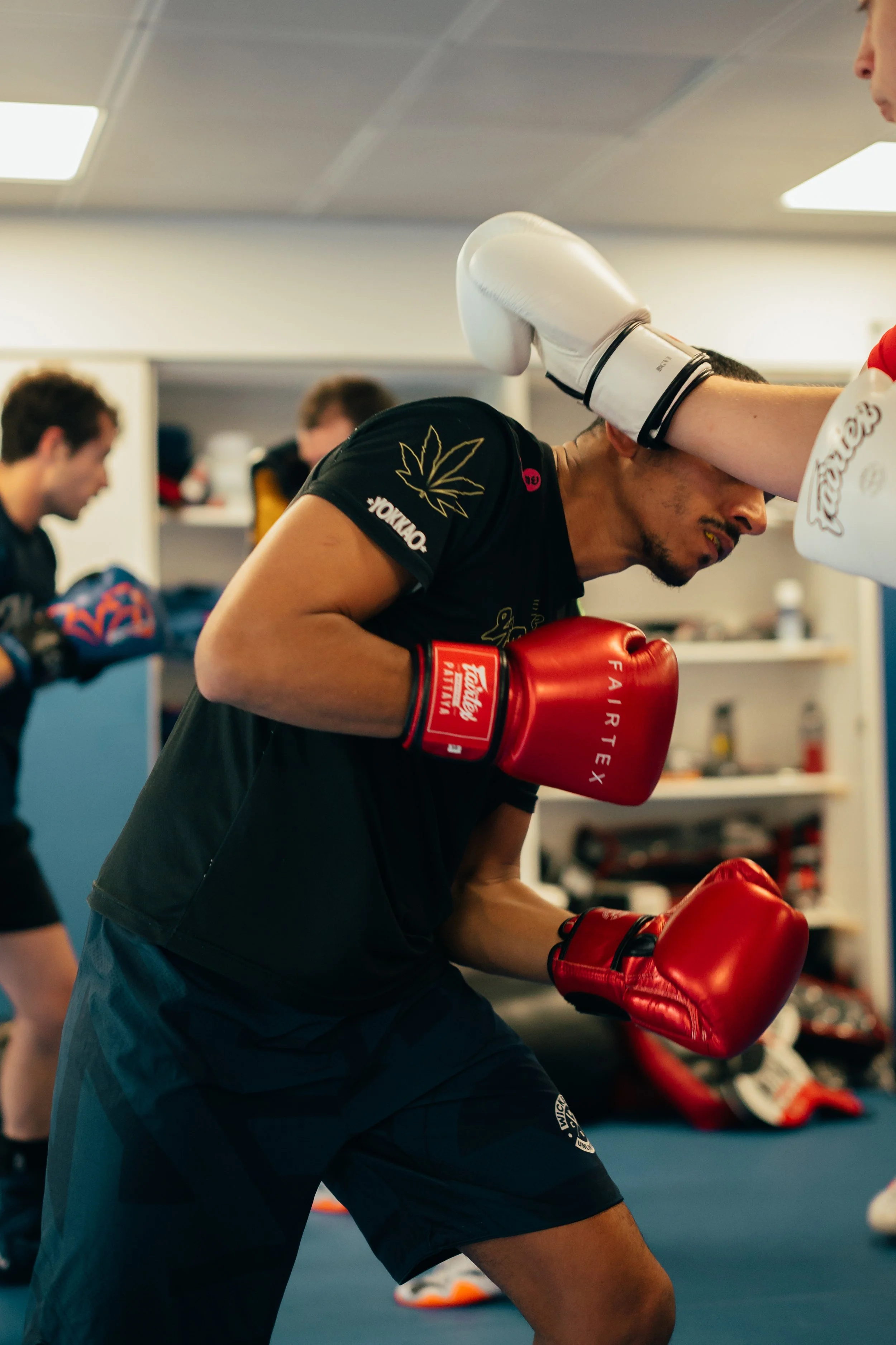 Un homme entraîné à la boxe s'entraîne dans un gymnase, portant des gants de boxe rouges et une tenue de sport noire. Il pratique sur un sac de frappe, avec d'autres personnes en arrière-plan