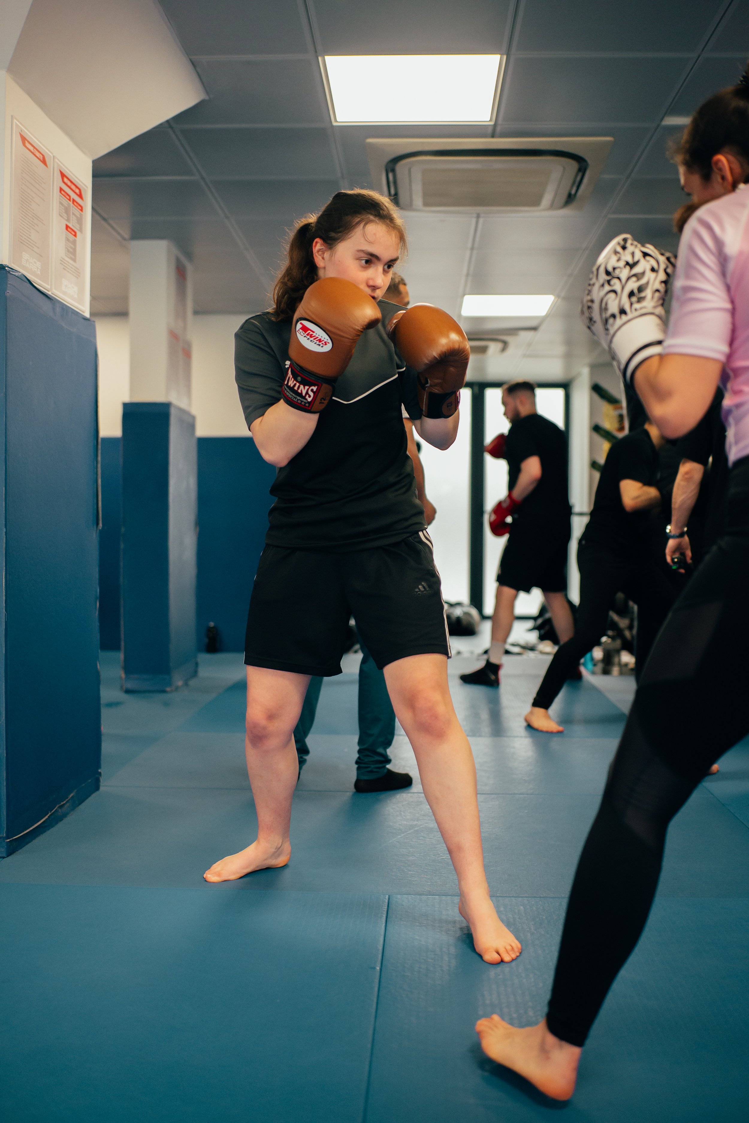 Une jeune fille en tenue de sport portant des gants de boxe marron en position de combat dans une salle de sport de boxe, avec d'autres personnes en arrière-plan, également engagées dans des activités d'entraînement.