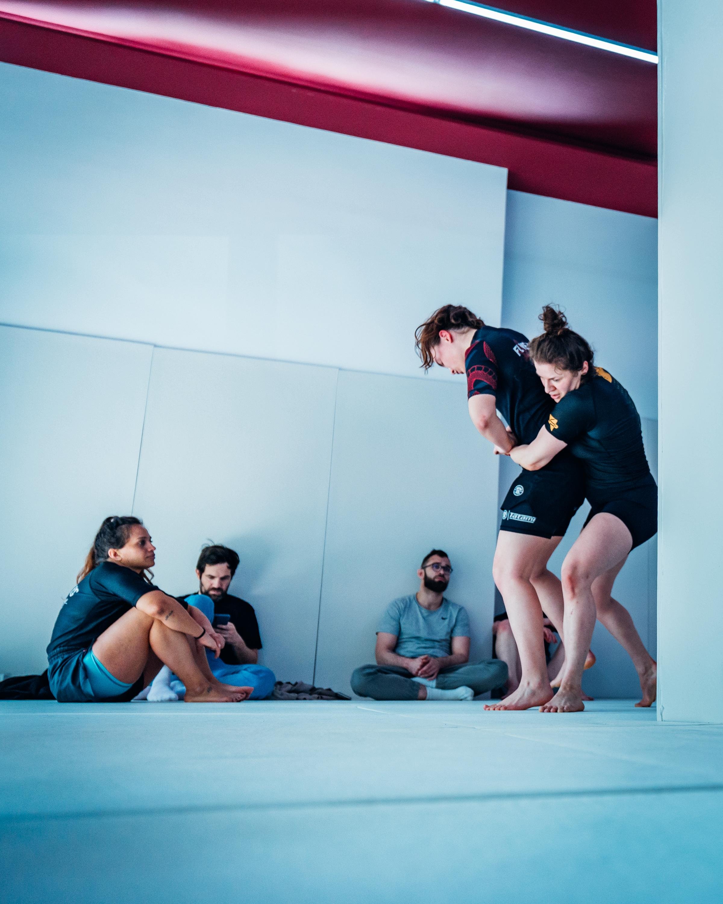Deux femmes en kimono de jiu-jitsu en train de s'entraîner, avec plusieurs personnes assises au sol dans un lieu d'entraînement.