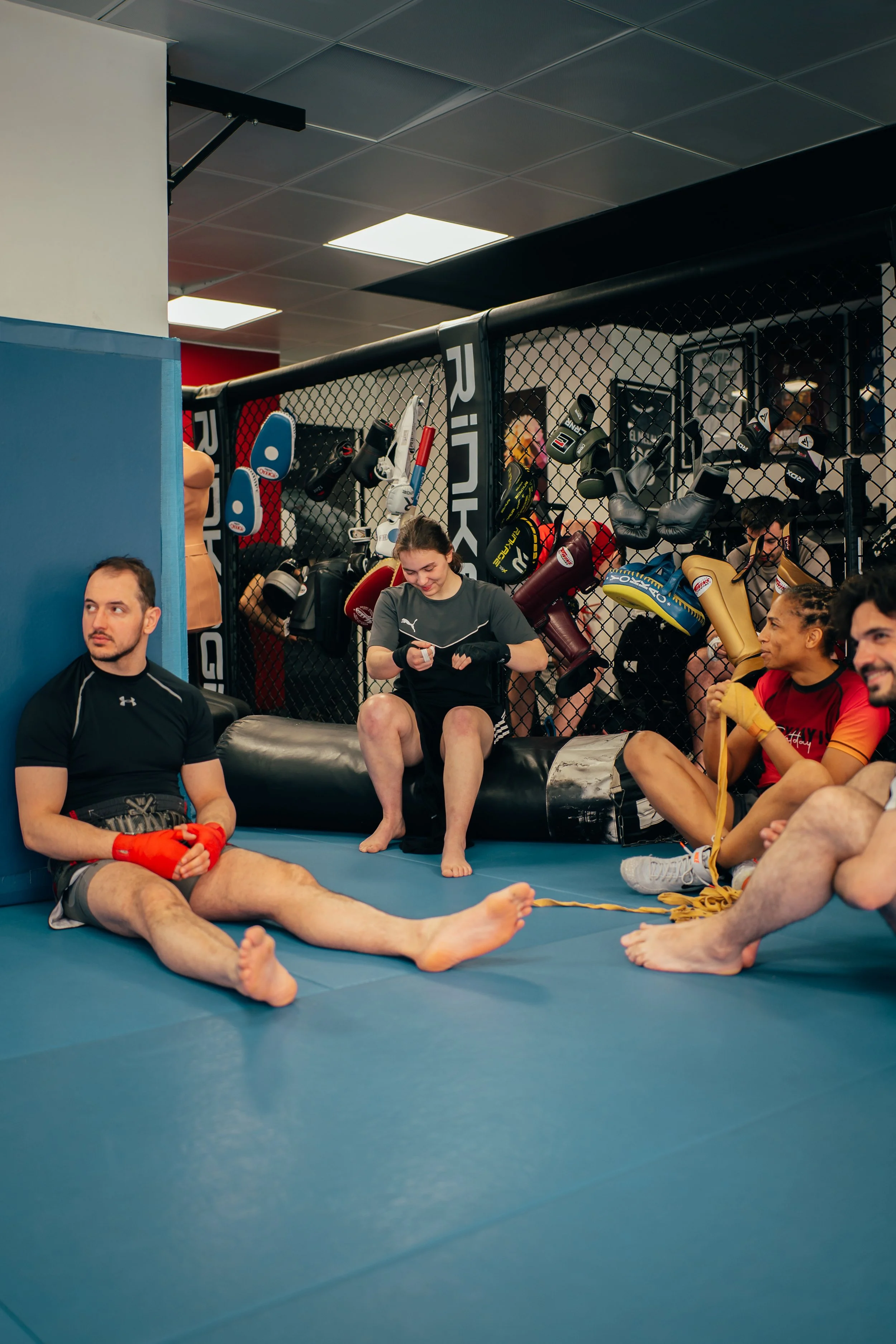 Des athlètes plusieurs boxeurs et boxeuses assis, un homme en position de repos sur le sol, un mur d'équipement de boxe en arrière-plan, après l'entraînement