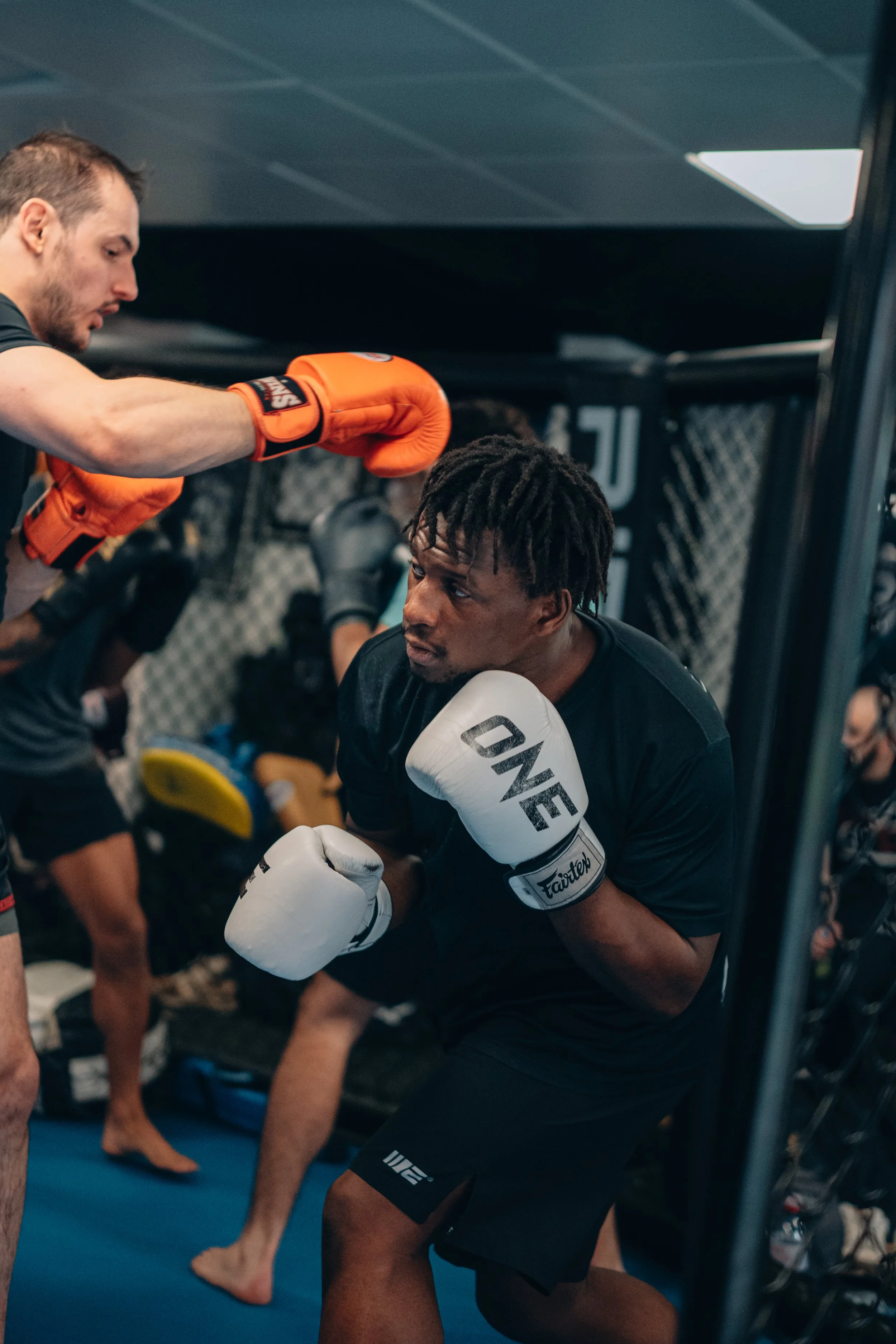 Deux hommes s'entraînent dans une salle de MMA, l'un en position de combat avec des gants de boxe, l'autre en préparation pour un entraînement, tous deux vêtus de vêtements de sport techniques.