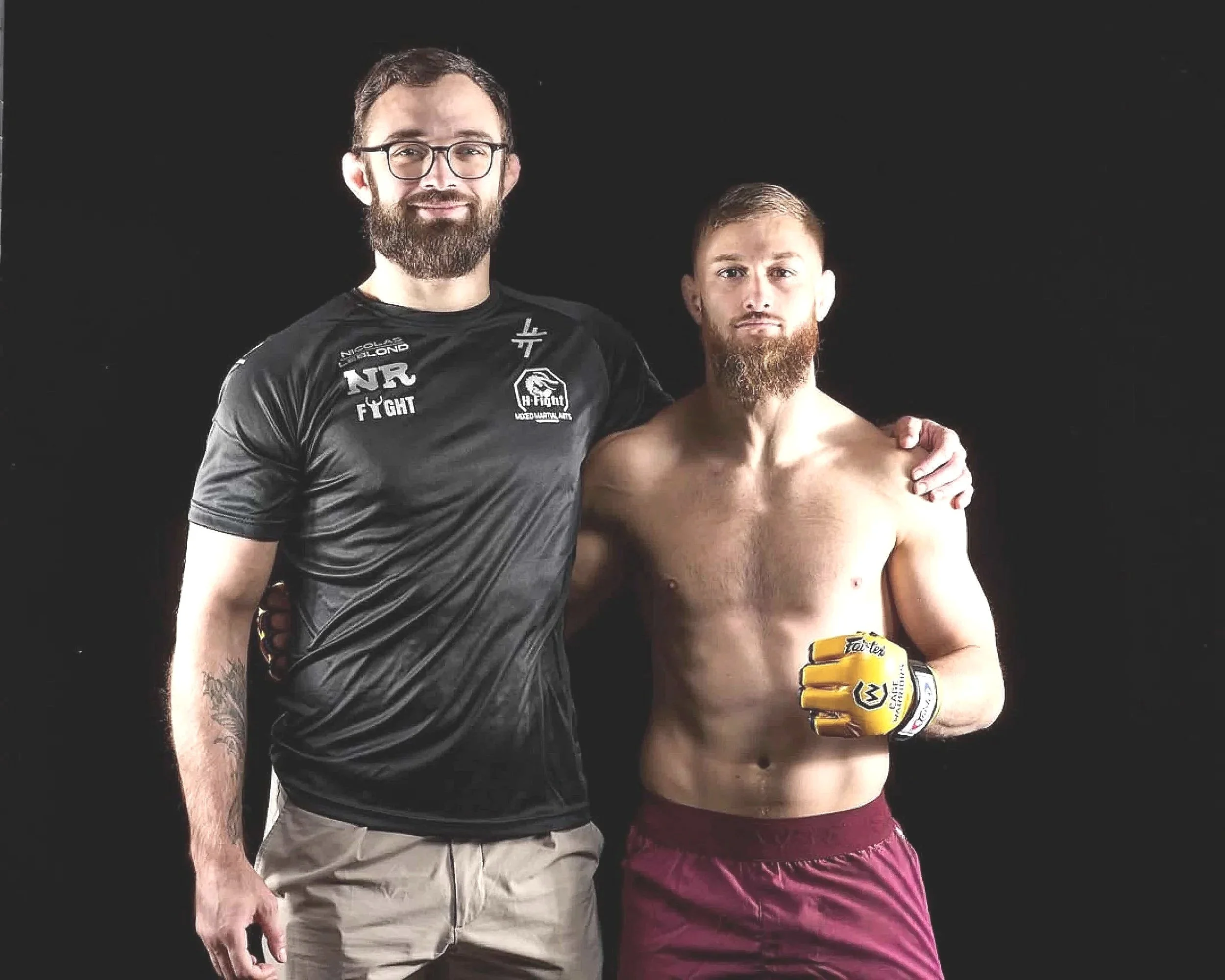 Deux hommes posent ensemble contre un fond noir, l'un portant un t-shirt de sport avec un logo et l'autre en short de combat, bras croisés, avec des gants de boxe jaunes. Ce sont des combattants professionnels de MMA