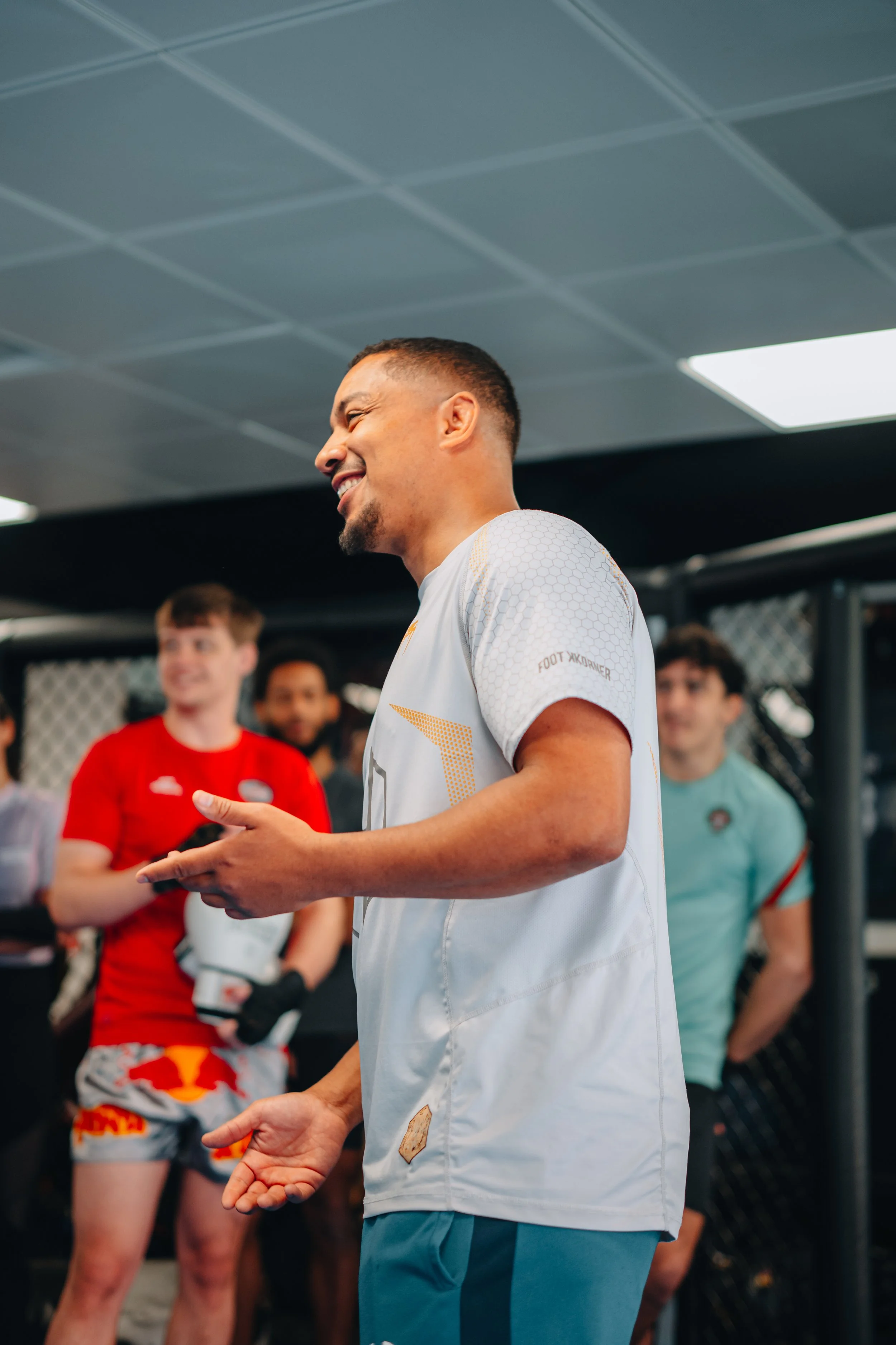 Homme souriant avec un tissu blanc dans une salle d'entraînement ou de sports, entouré de jeunes hommes et femmes.
