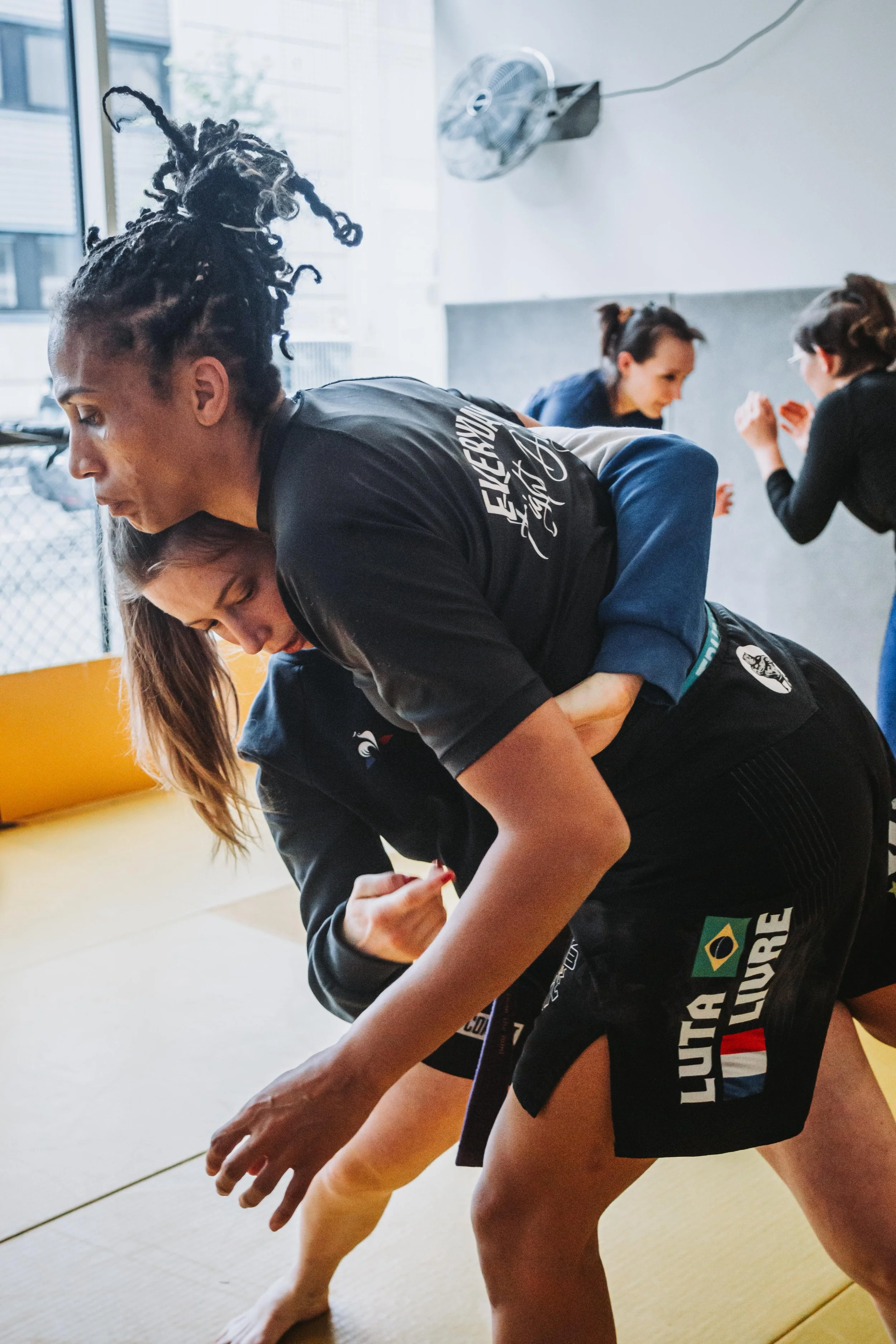 Deux femmes pratiquant la lutte dans une salle d'entraînement.  Elles s'engagent dans un combat au sol sur un tapis jaune avec une fenêtre derrière elles.