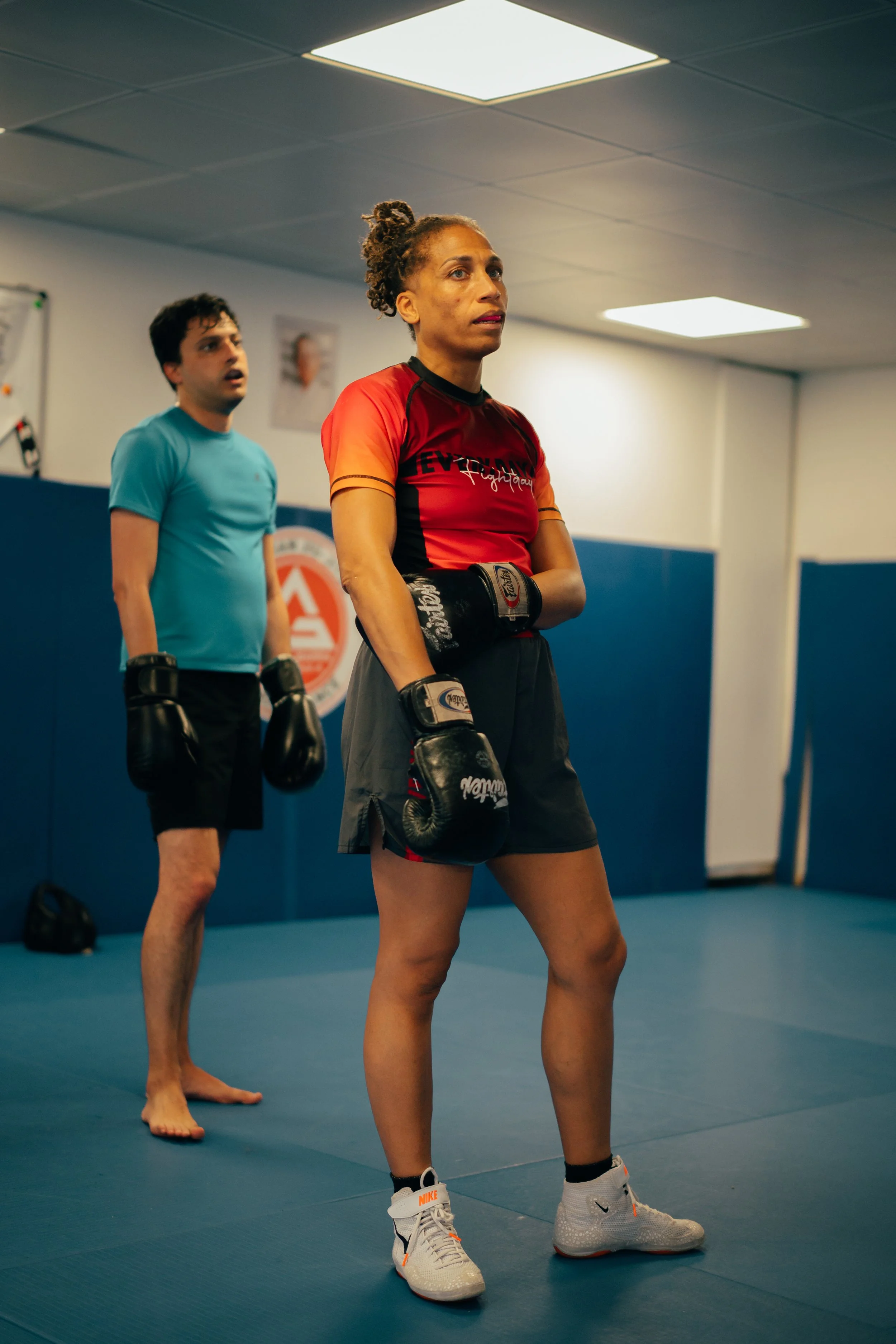 Deux personnes pratiquent la boxe dans une salle d'entraînement. La femme en avant porte des gants de boxe, une robe de sport rouge et grise, et des chaussures de sport Nike. La personne à l'arrière porte un t-shirt bleu et des gants de boxe, et semb