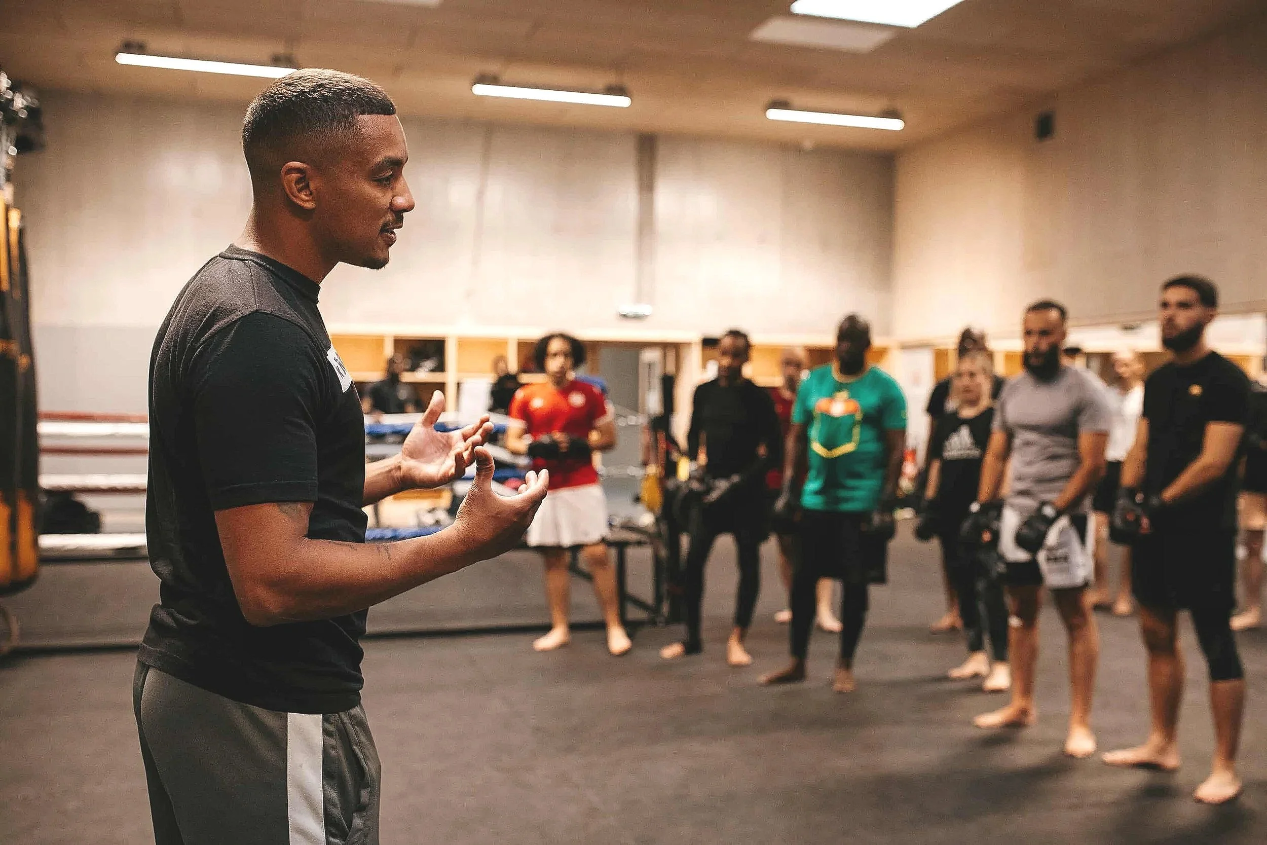 Un entraineur de MMA en train de parler à un groupe de sportifs et sportives en tenue de sport dans une salle d'entraînement.