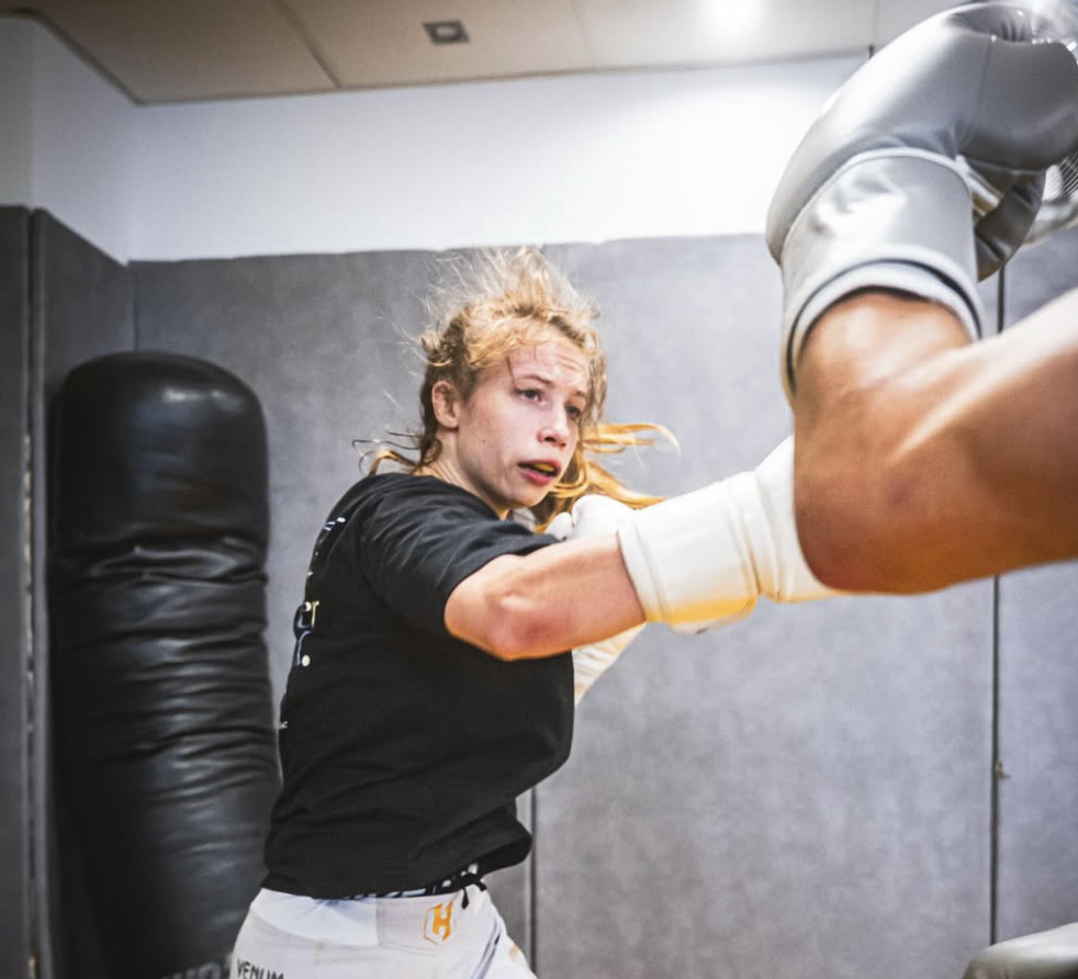 Une femme en équipement de boxe, portant des gants et un short, esquive un coup d'une personne qui porte des gants de boxe, dans une salle d'entraînement.