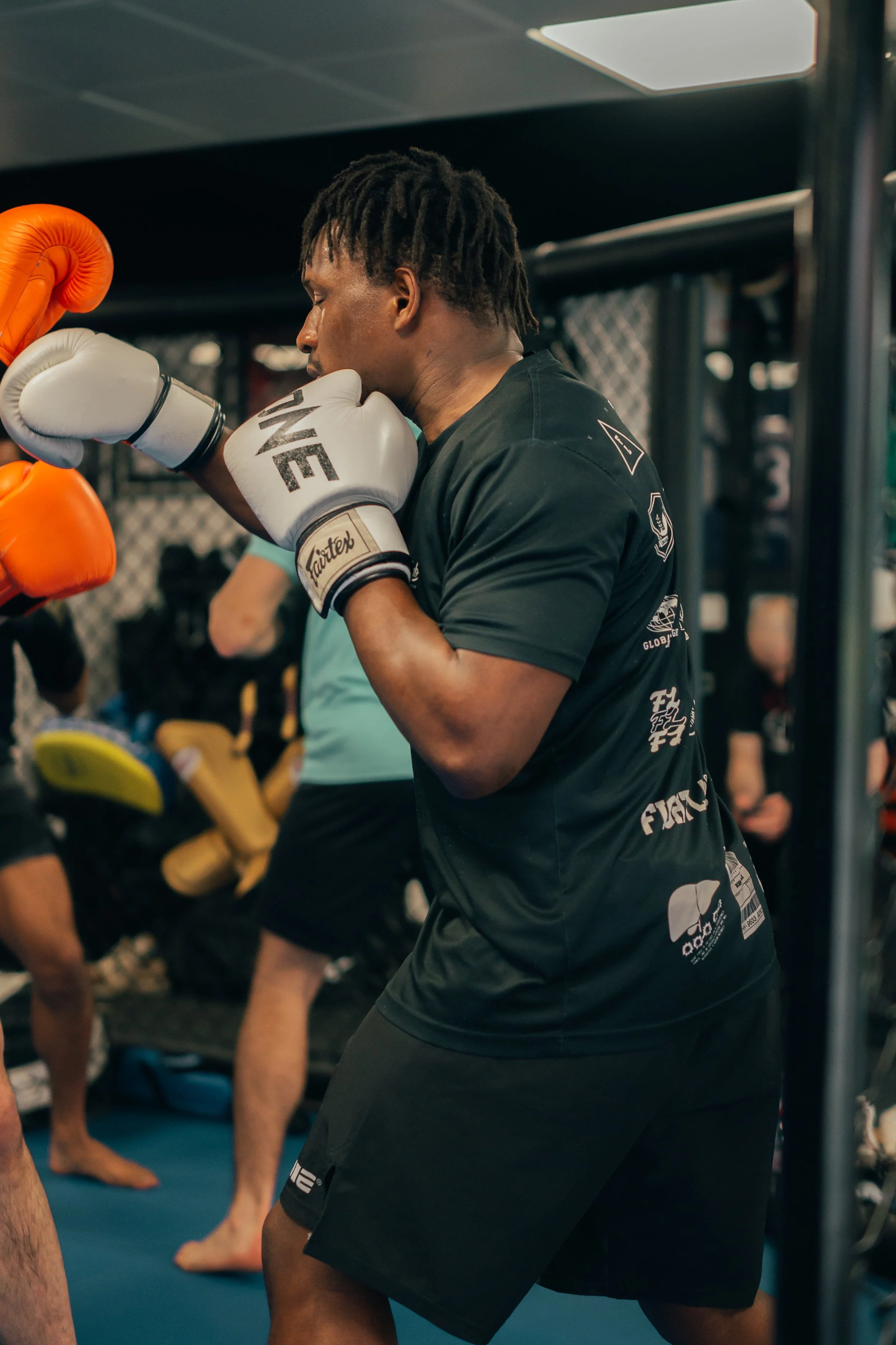 Un homme pratique la boxe dans une salle d'entraînement, portant des gants de boxe blancs et noirs, concentré, avec d'autres personnes visibles en arrière-plan.