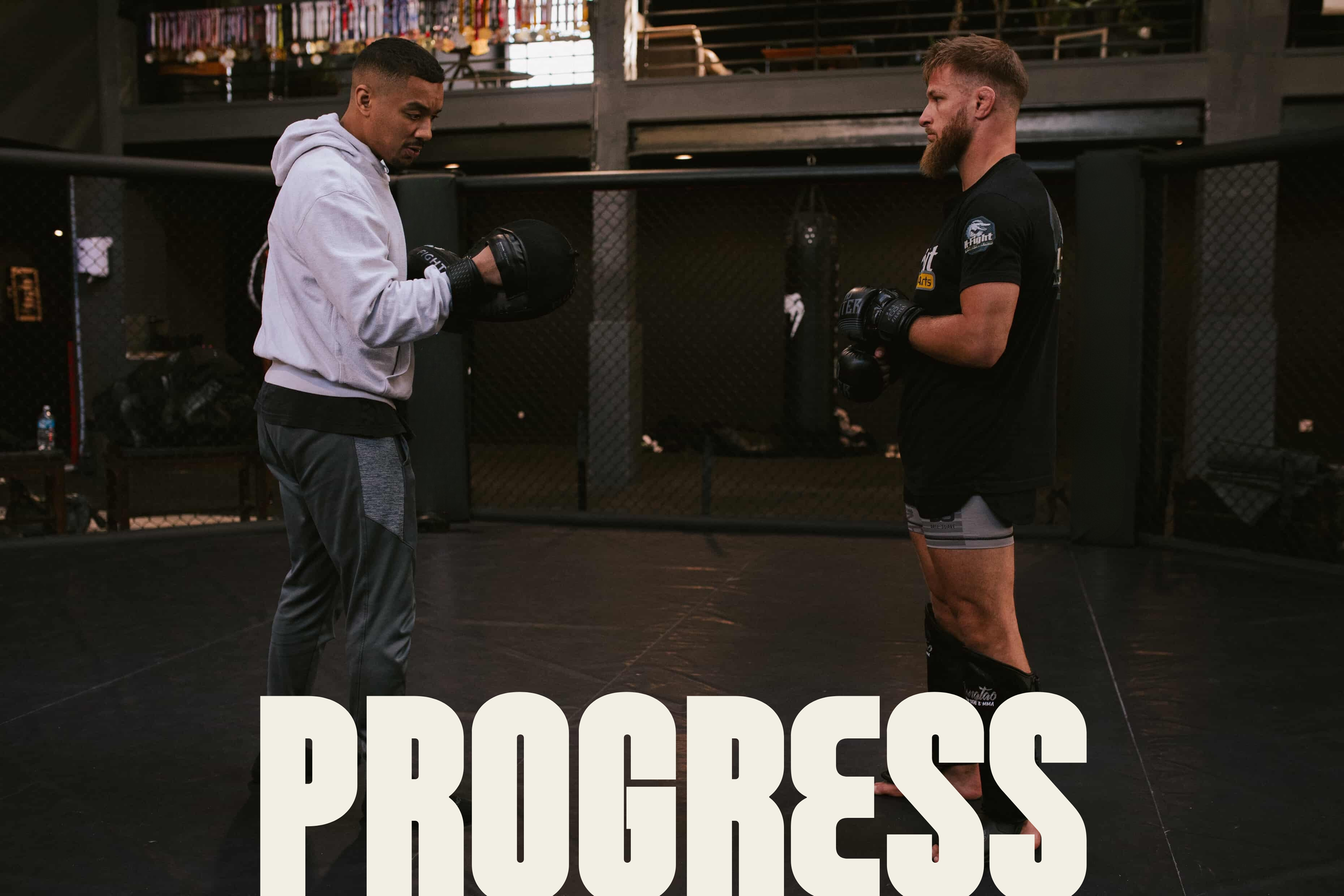 Deux hommes en tenue de combat de MMA dans un gymnase, se regardant en se tenant en position de combat, avec un panneau écrivant 'PROGRESS' partiellement visible en bas.