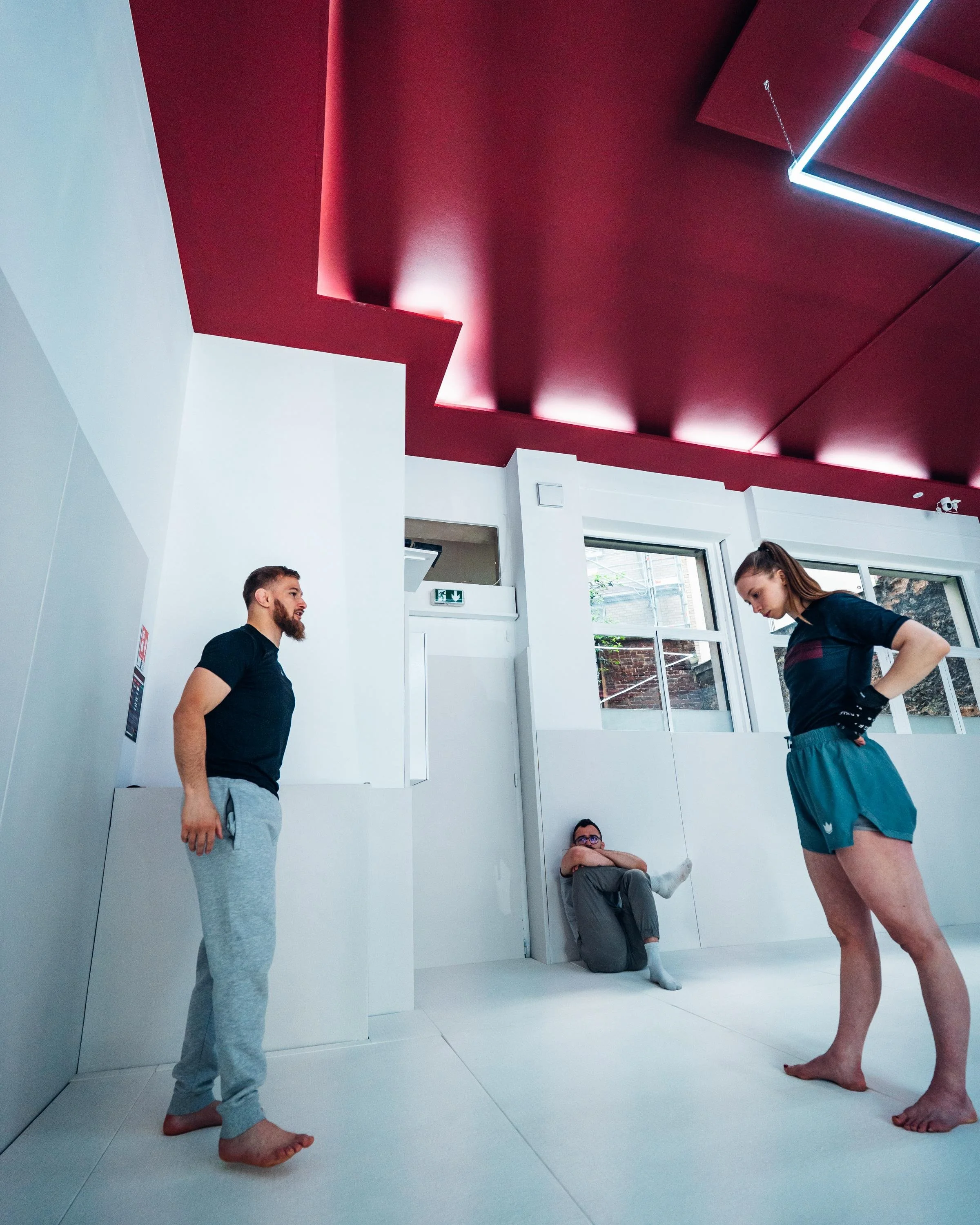 Deux personnes dans une salle blanche, l'une debout et l'autre assise contre le mur, le regard vers le sol. Un troisième individu, féminin, se tient en face, les mains sur les hanches, regardant vers le bas.