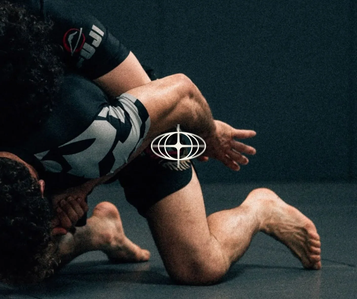 Deux hommes s'entraînent en jiu-jitsu brésilien sur un tapis noir, avec un symbole blanc au centre de leur contact.