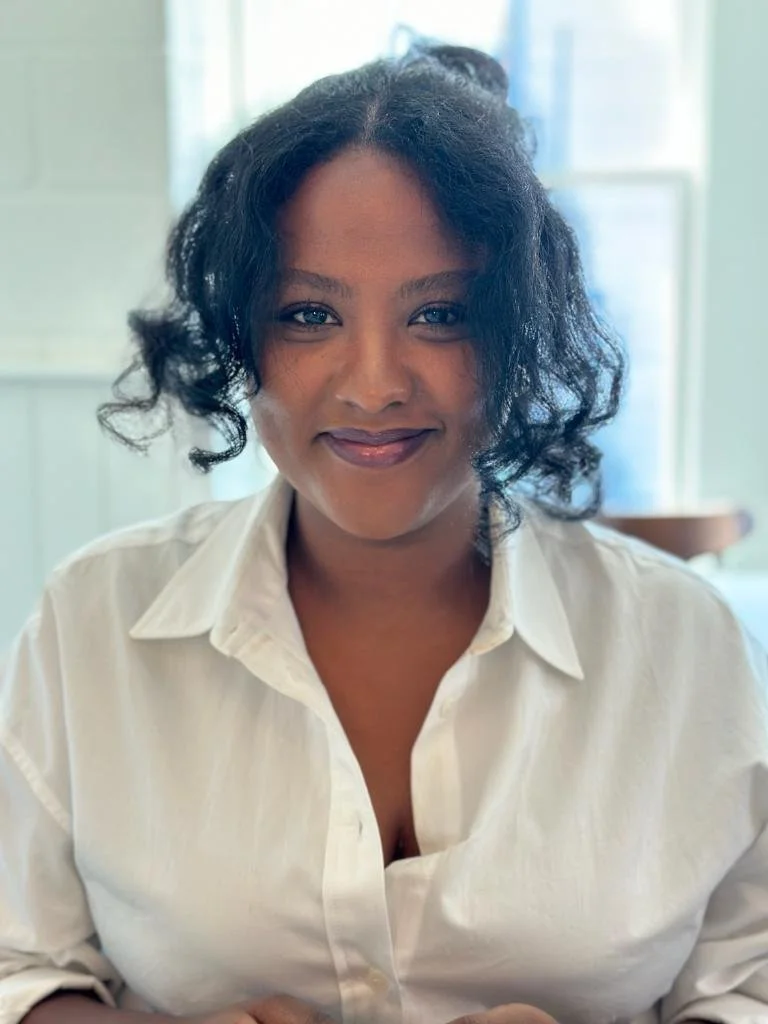 A woman with short, curly black hair wearing a white button-up shirt, smiling softly at the camera, in a well-lit room with a window in the background.