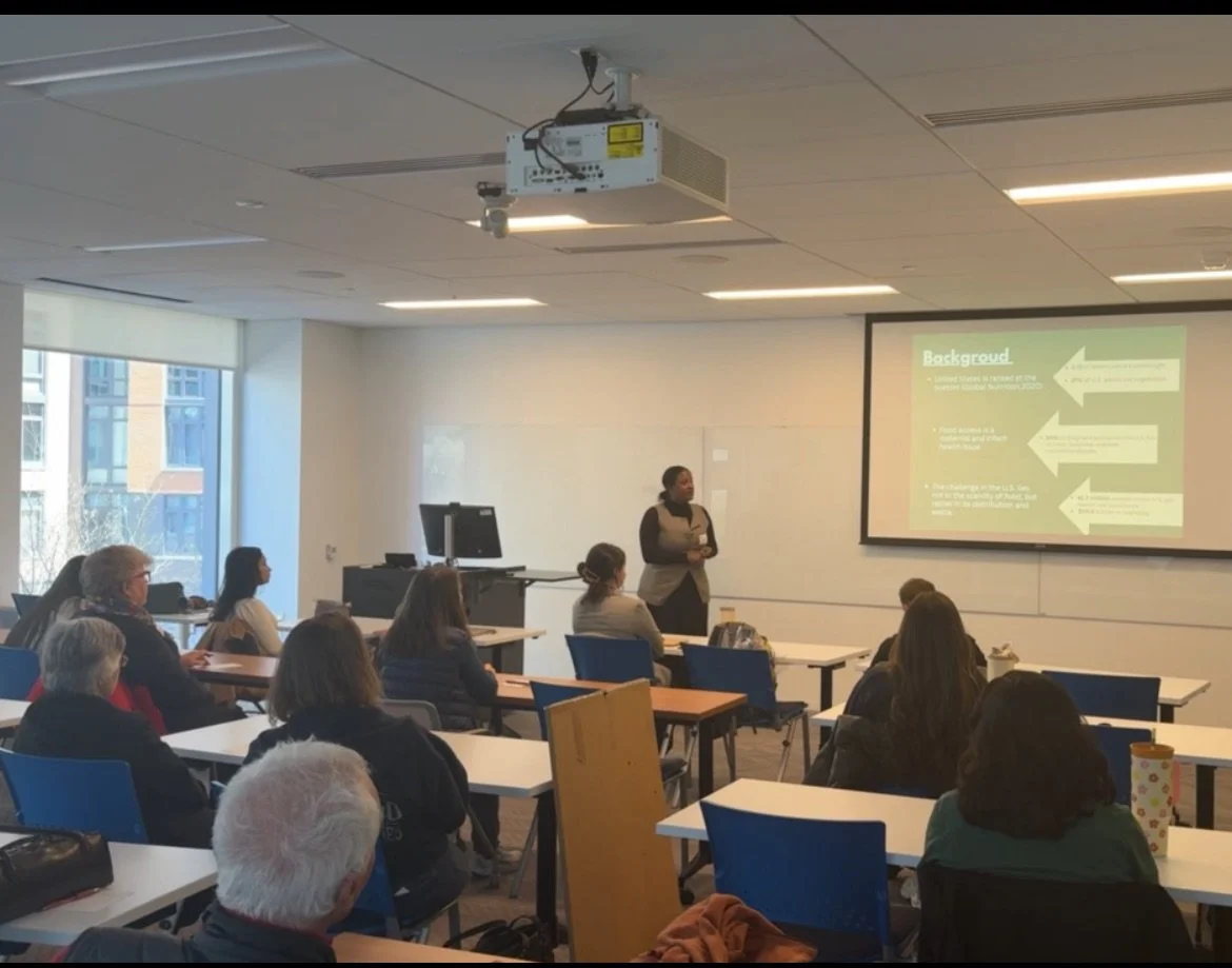 A woman gives a presentation in a classroom with seated adult attendees, a projector, and a large screen displaying a presentation slide titled 'Background'. Large windows behind the audience provide natural light.