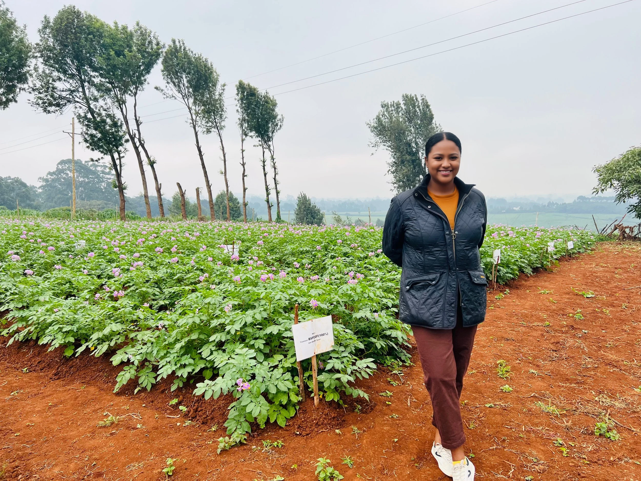A woman standing in a flower field on a farm, with trees and power lines in the background, smiling at the camera.