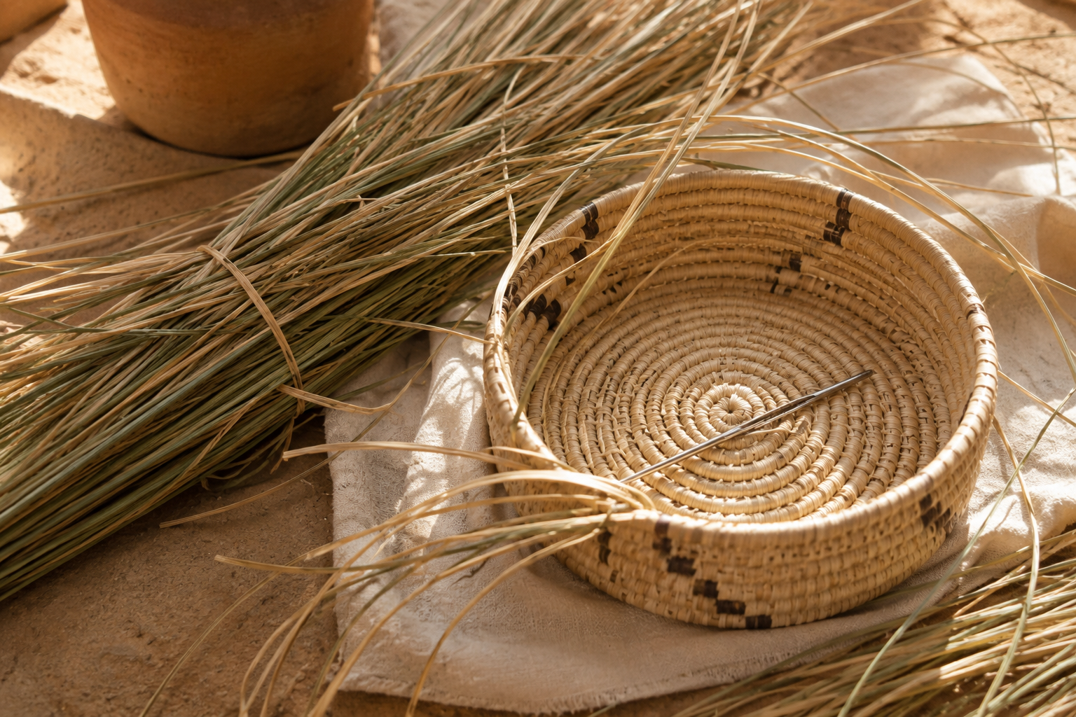 A woven basket with a metal knitting needle inside, surrounded by dried grass and a piece of cloth, set on a sandy surface.