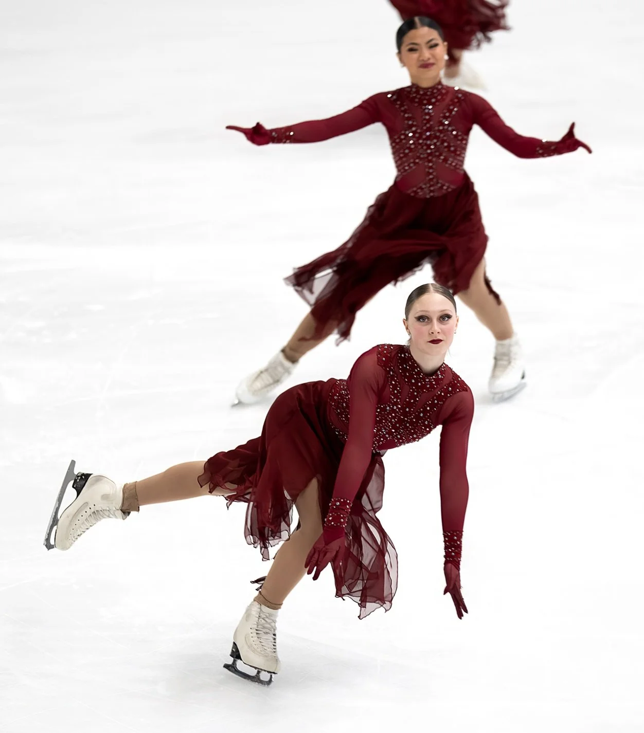 Two female figure skaters in matching burgundy dresses with embellishments performing on ice, with one skating and the other in a low pose on the ice.
