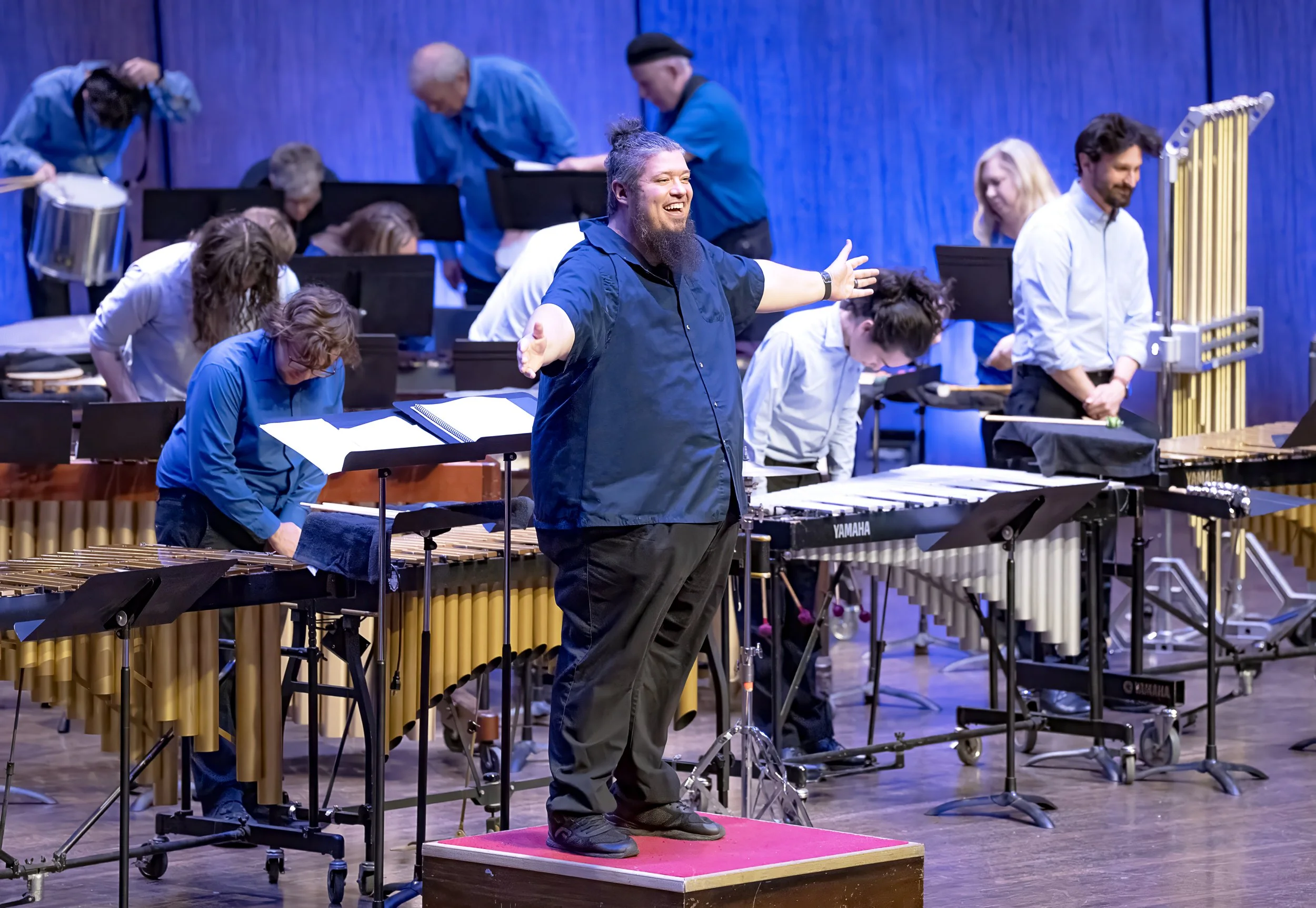 A conductor standing on a red platform leading a music ensemble, with musicians playing percussion and wind instruments on a stage with a blue backdrop.