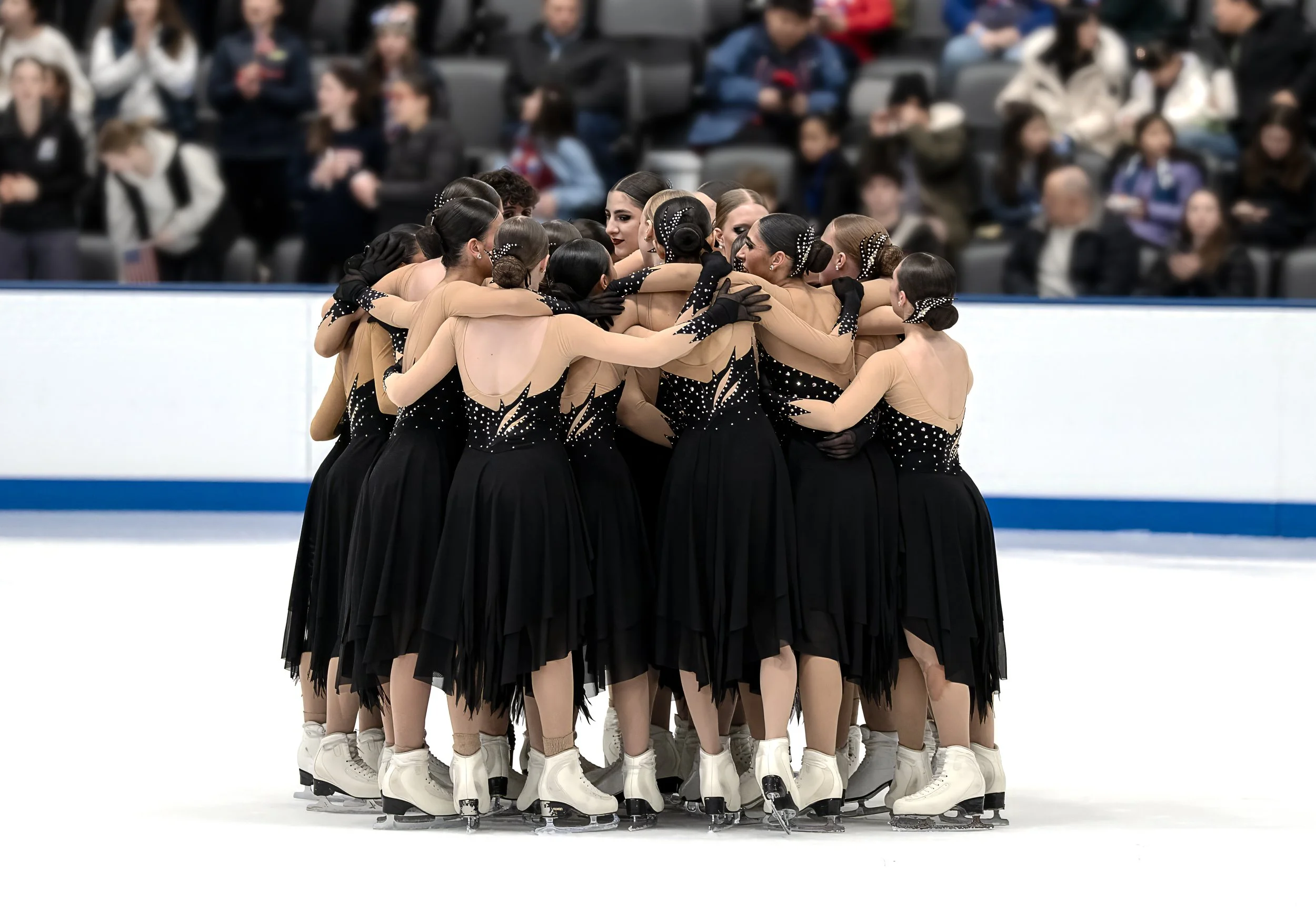 A group of female figure skaters in tan and black costumes huddle together before a performance on the ice rink.