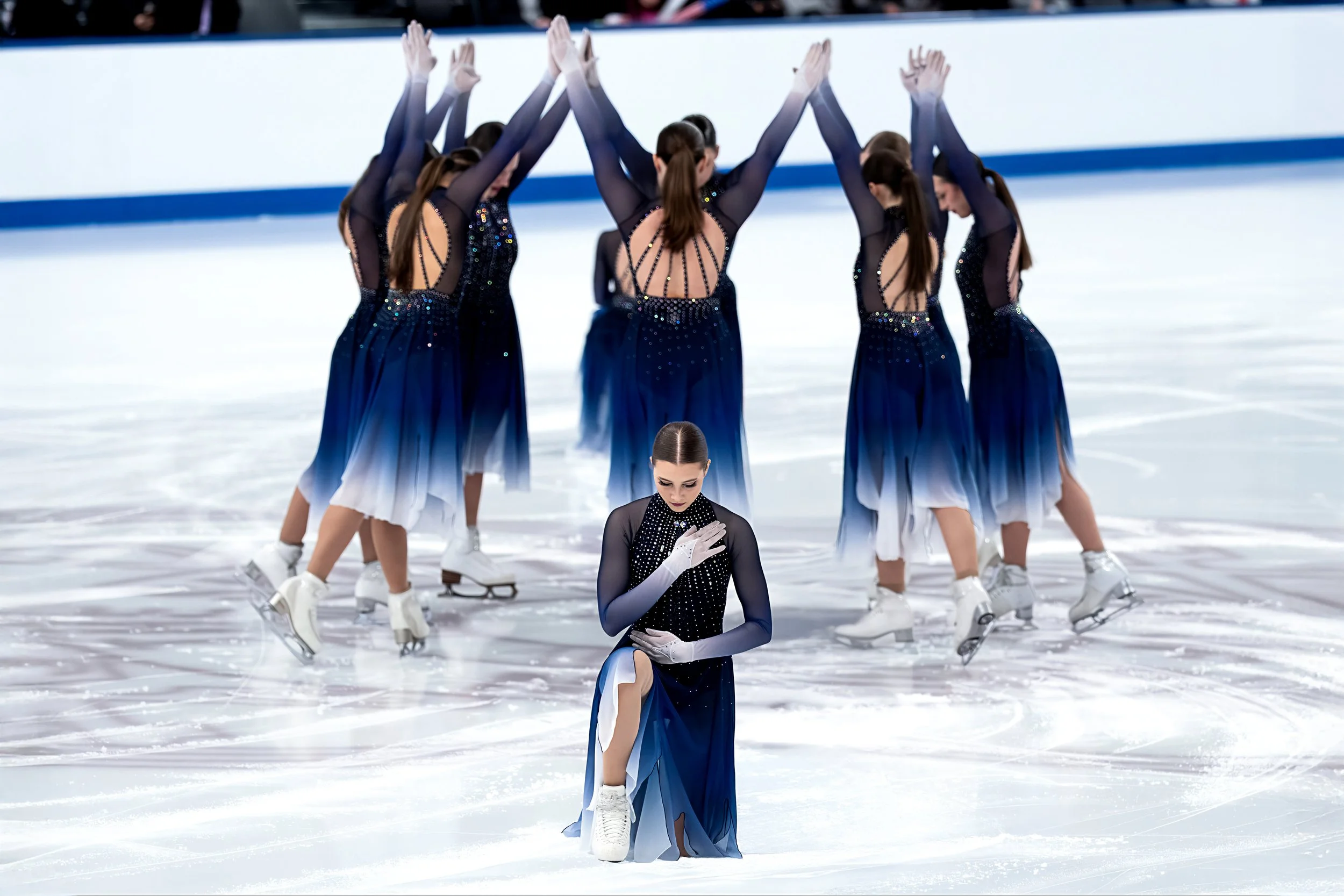 Figure skaters wearing coordinated black and blue costumes performing a routine on ice, with one skater kneeling in front and others raising their hands in a group pose.