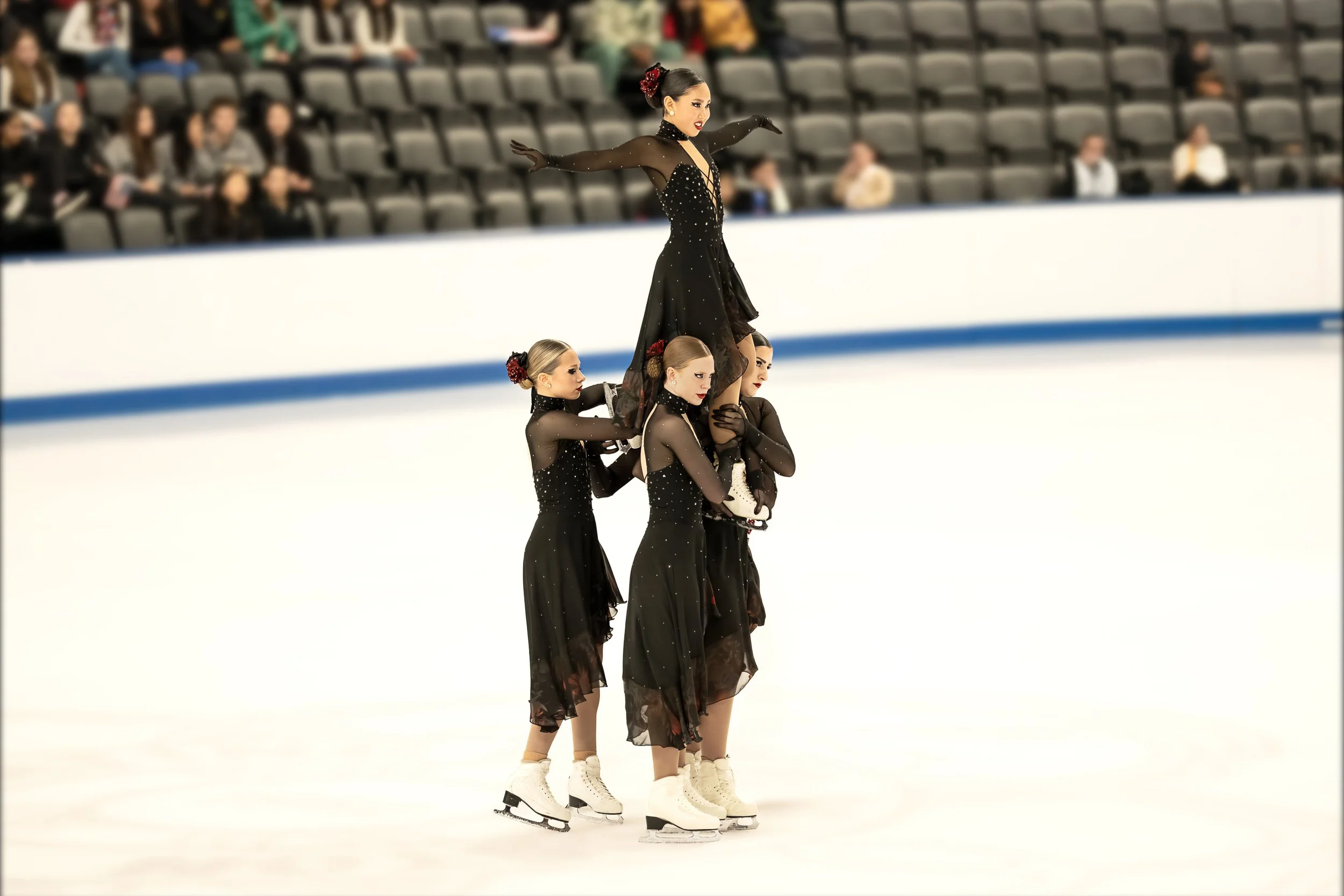 Four female figure skaters in black costumes with rhinestones, one standing on the skaters' shoulders, performing a routine on the ice rink during a competition or exhibition.