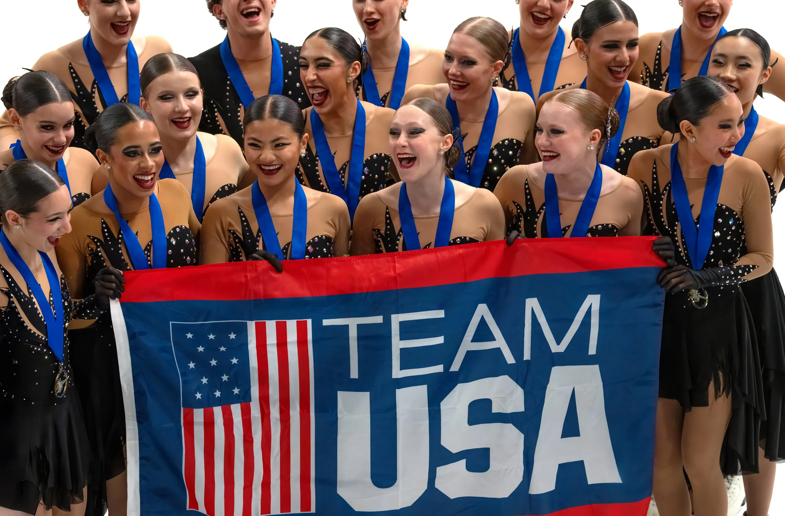 Group of female gymnasts celebrating after a competition, holding a banner that reads 'TEAM USA', wearing black and gold leotards with rhinestone embellishments, each with a blue medal around their neck.